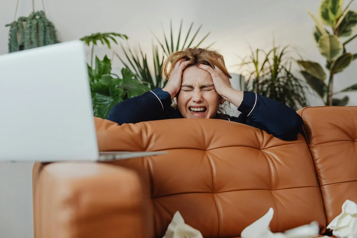 A woman sitting on a brown leather couch, holding her head with both hands, appearing stressed or frustrated. She has red hair and is wearing a dark long-sleeve top. There are green plants in the background and a laptop on the armrest of the couch.