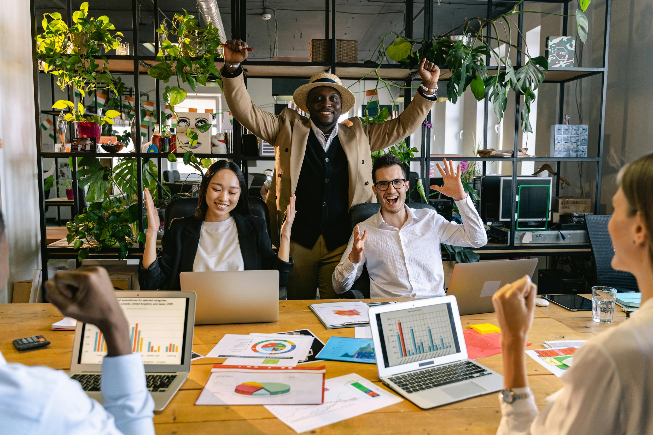 A diverse group of people in an office, with one man standing and raising his arms, surrounded by colleagues seated at a table with laptops and documents, celebrating or presenting.