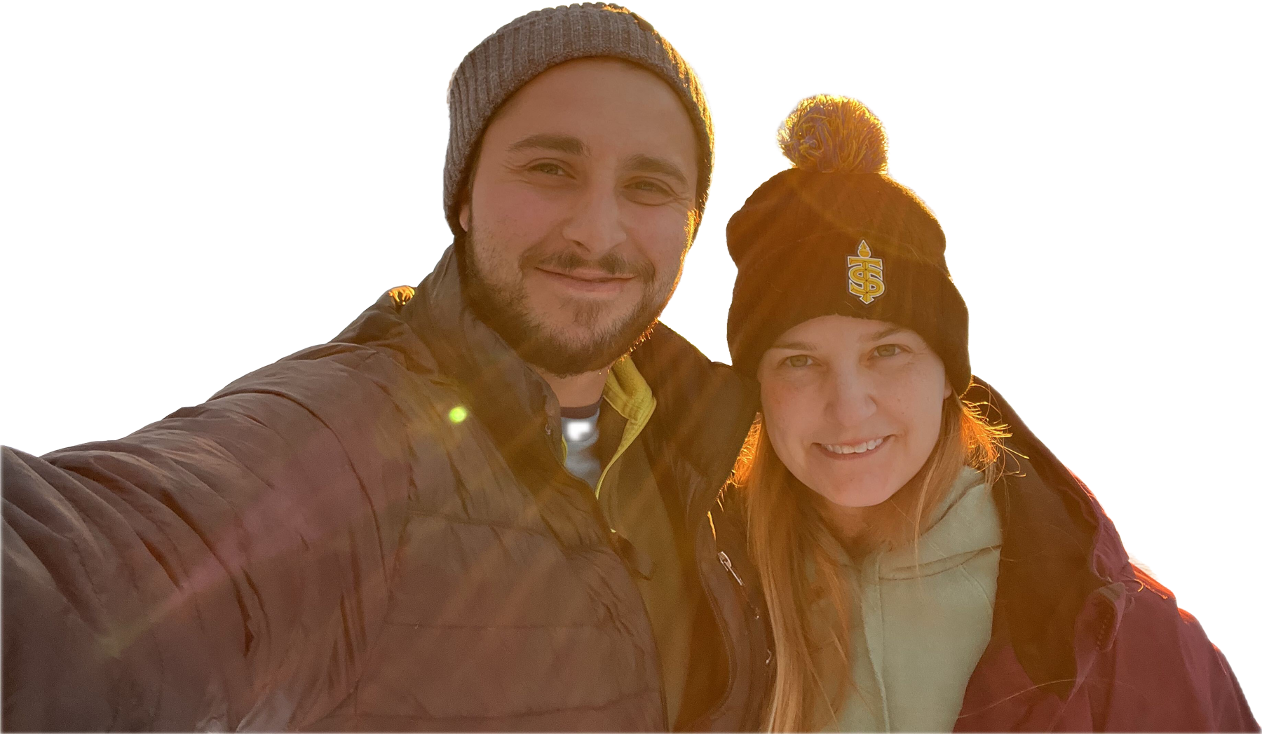 A smiling man and woman taking a selfie outdoors during sunset, both wearing warm clothing and hats.