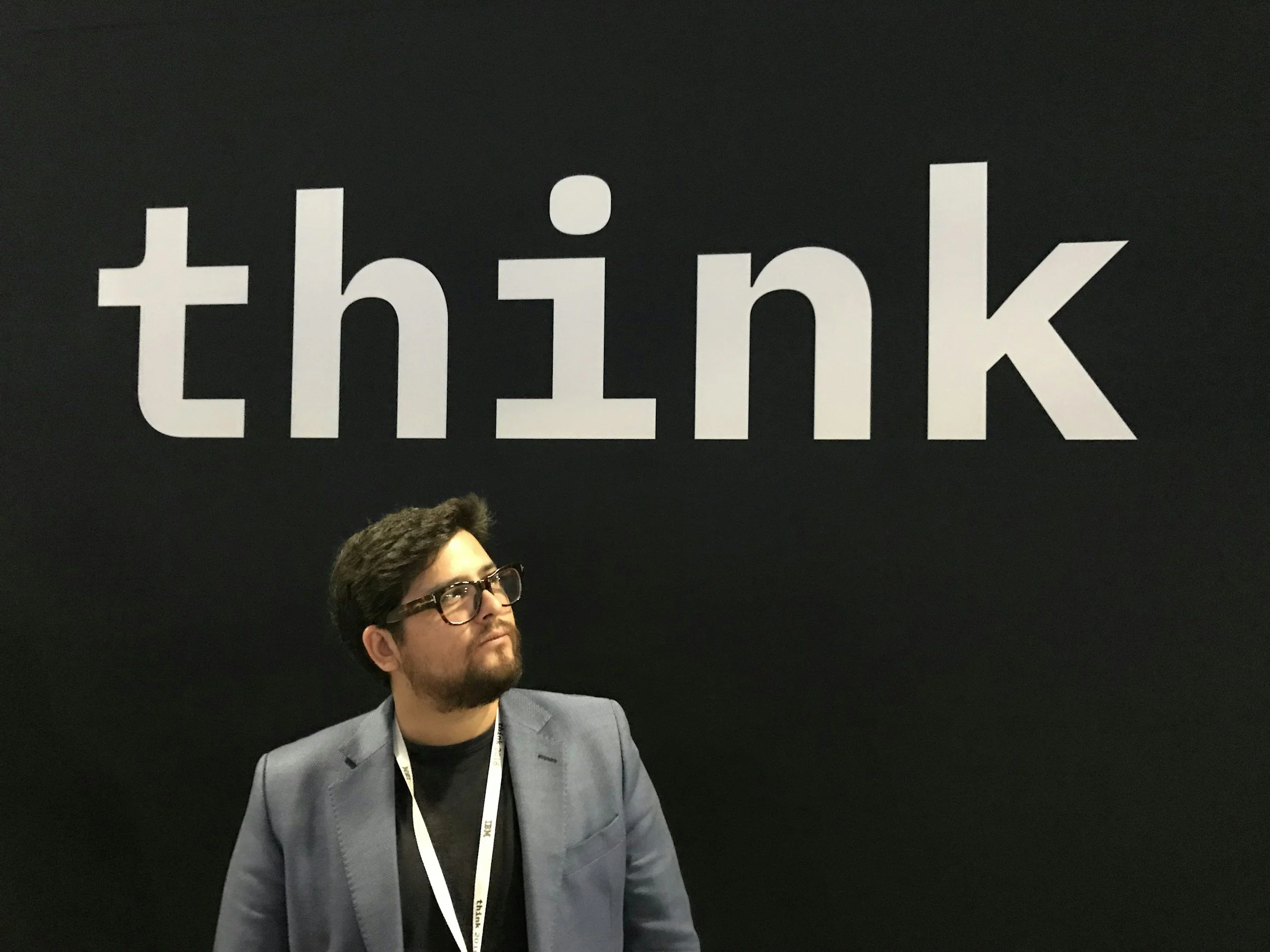 A man with dark hair, glasses, and a beard, dressed in a gray suit and black shirt, is standing in front of a black background with the word 'think' in large white lowercase letters.