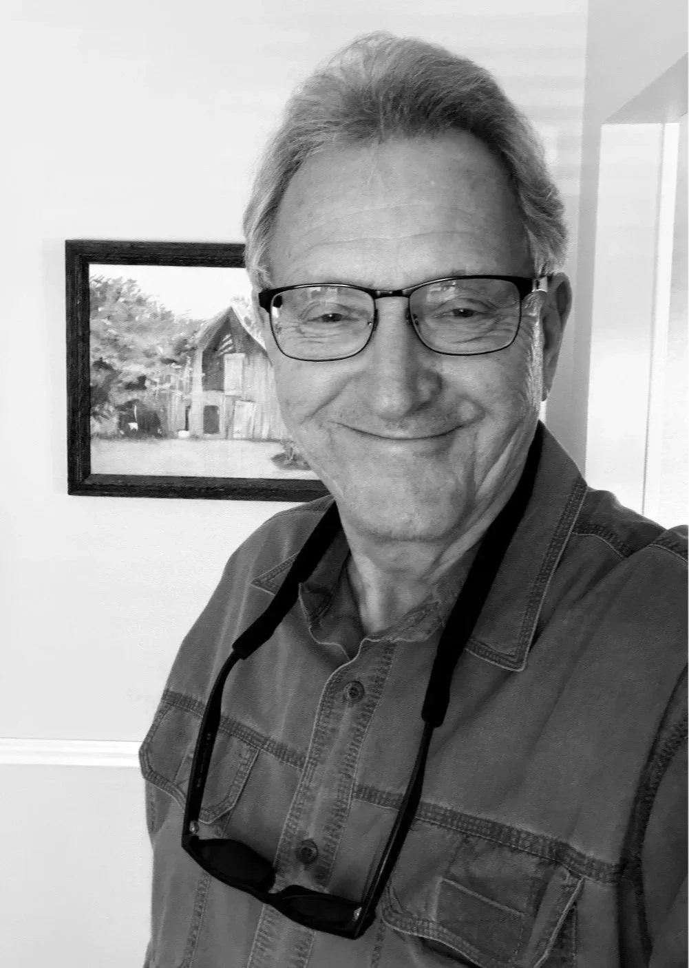An older man with glasses and gray hair taking a selfie indoors, with a framed painting of a barn and trees hanging on the wall behind him.