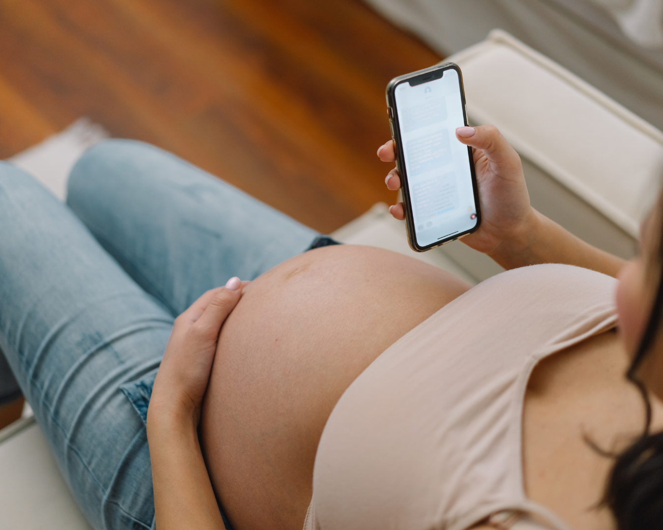 Schwangere Frau sitzt auf einem Stuhl und liest eine Nachricht auf ihrem Smartphone, während sie ihre Hand auf ihren Babybauch legt.