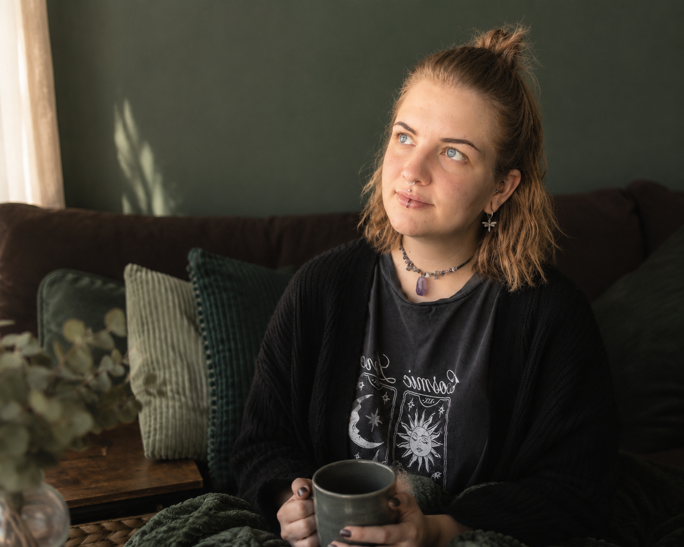 Eine junge Frau mit blonden, leicht gewellten Haaren, die nach oben frisiert sind, sitzt auf einem Sofa vor einer grünen Wand. Sie trägt ein schwarzes T-Shirt mit einem mystischen Design und einer schlichten schwarzen Strickjacke. Sie hält eine graue Tasse in beiden Händen und schaut nach oben, während sie an etwas denkt. Im Hintergrund sind mehrere bunte Kissen, und die Szene wirkt gemütlich und entspannt.
