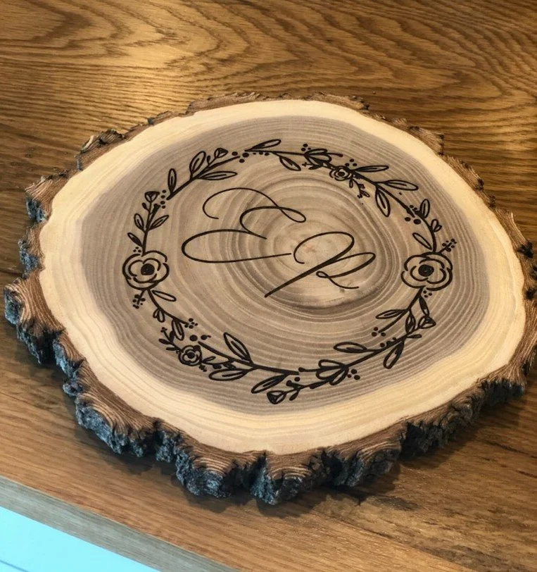 A wooden slice with engraved decorative floral wreath and the initials 'E & M' in the center.