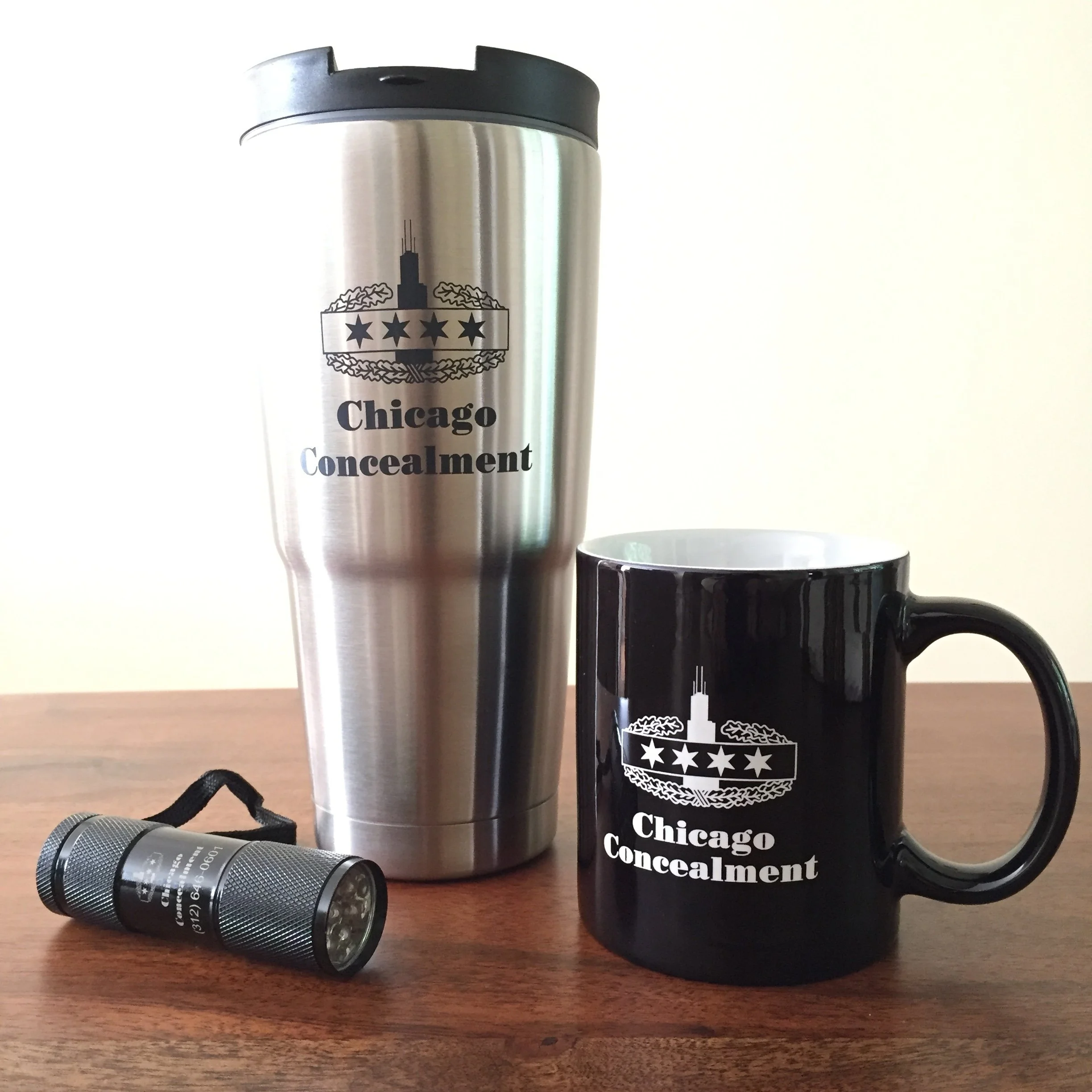 A stainless steel travel tumbler with a black lid, a black mug, and a small flashlight on a wooden surface. All items display the Chicago Conference logo and text.