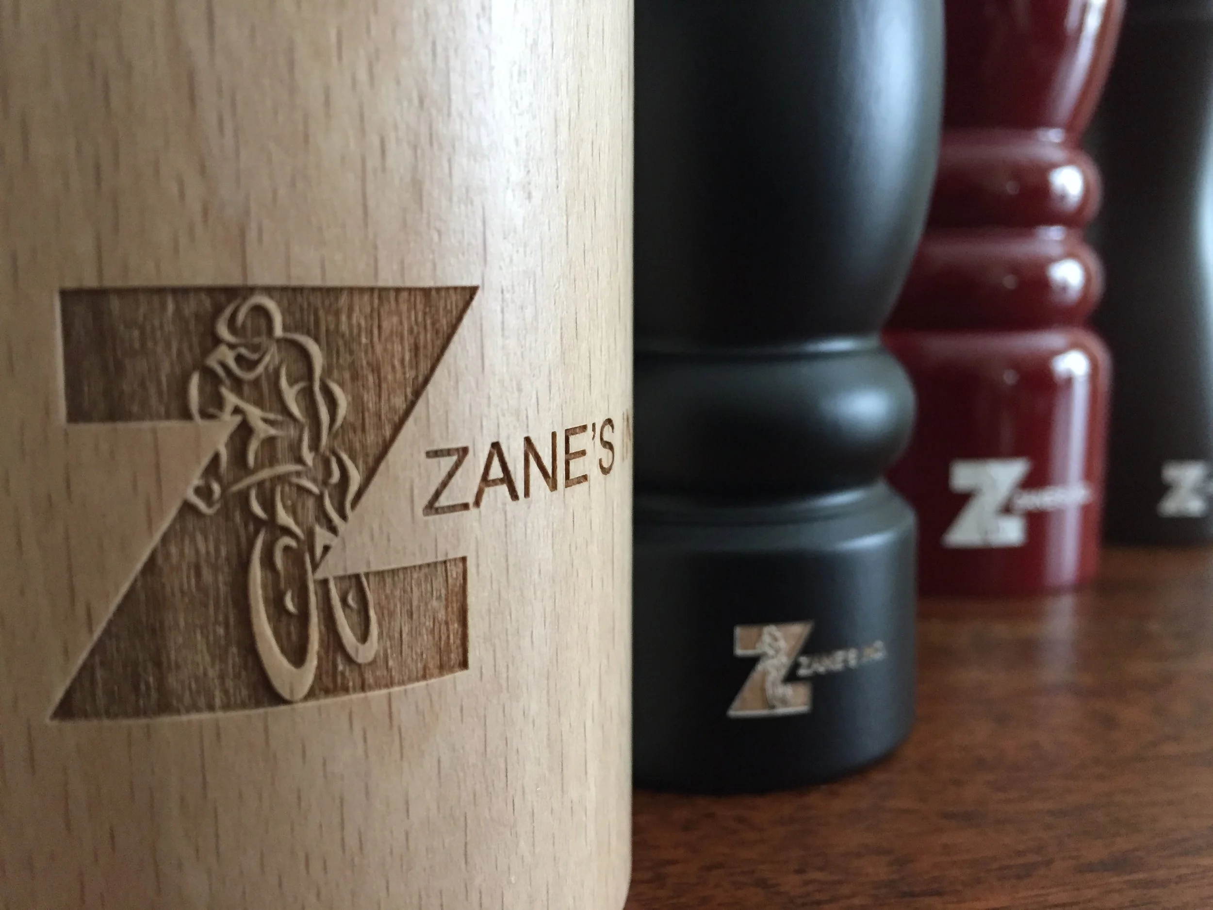 Close-up of a wooden Zane's box with an engraved chef logo on the lid. Behind the box, there are stacked black and red containers with white text and numbers.