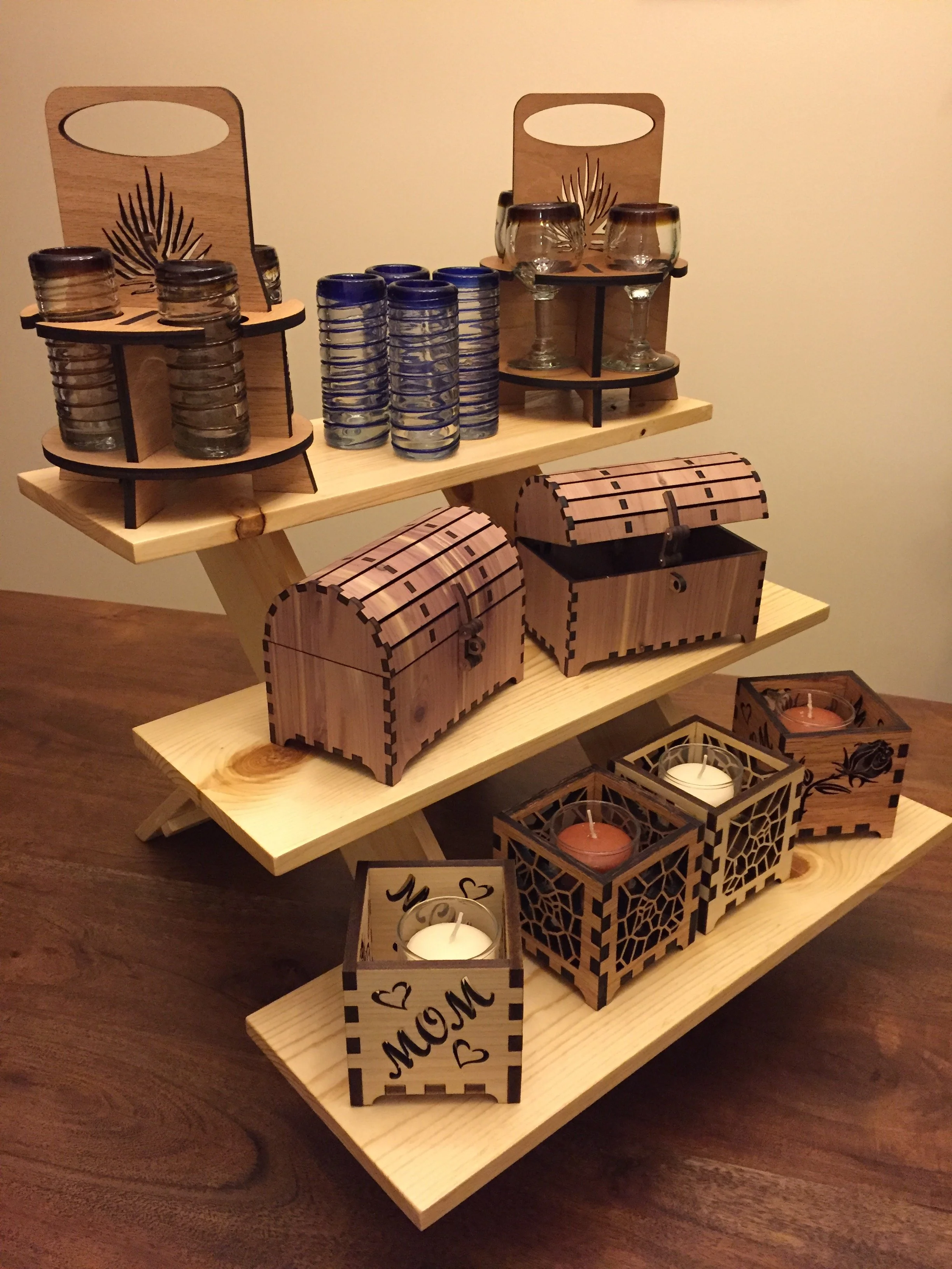 Three wooden tiered shelves display various candles and glassware, with decorative wooden boxes and lanterns at the bottom, against a plain wall.