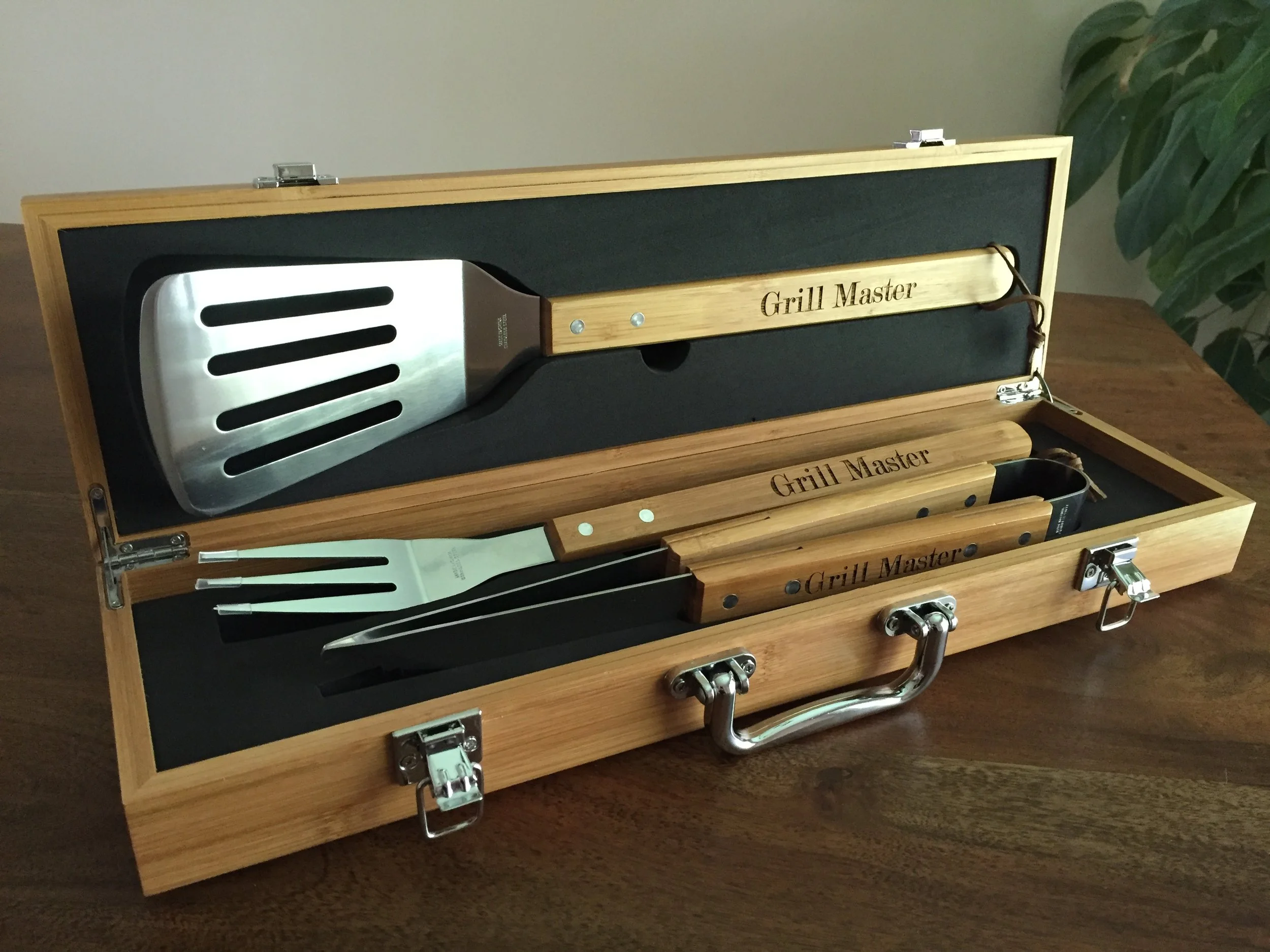 A wooden box labeled "Grill Master" containing metal grilling tools, including a spatula and tongs, with a metal handle on each tool, resting on a wooden surface next to a large green leafy plant.