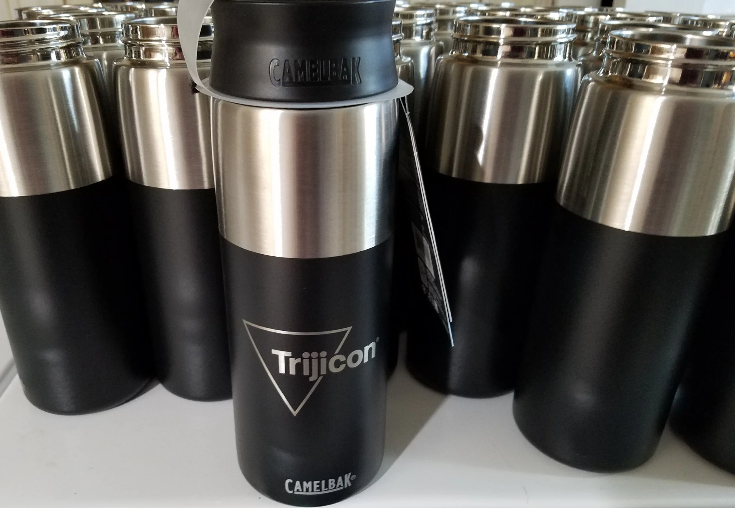 Black stainless steel travel mug with a black lid, labeled 'Camelbak Trijicon,' among similar mugs on a white surface.