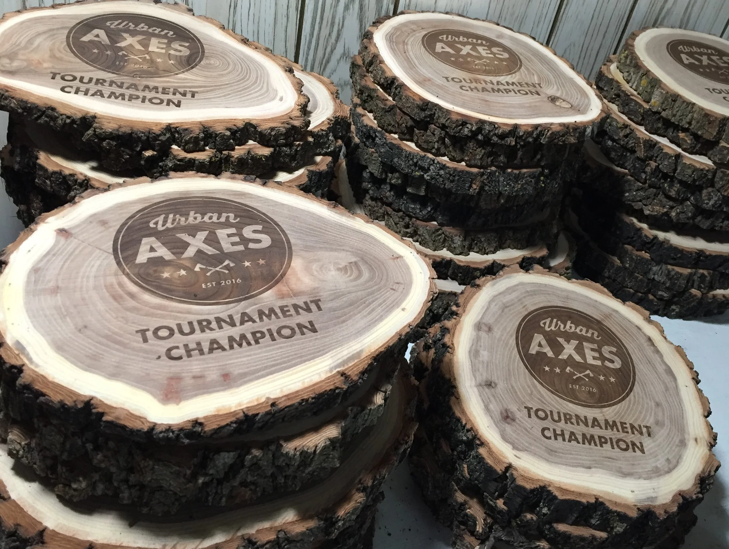 Stacks of round wooden plaques with bark edges and printed logos for the 'Urban Axes' tournament champion, established 2016.
