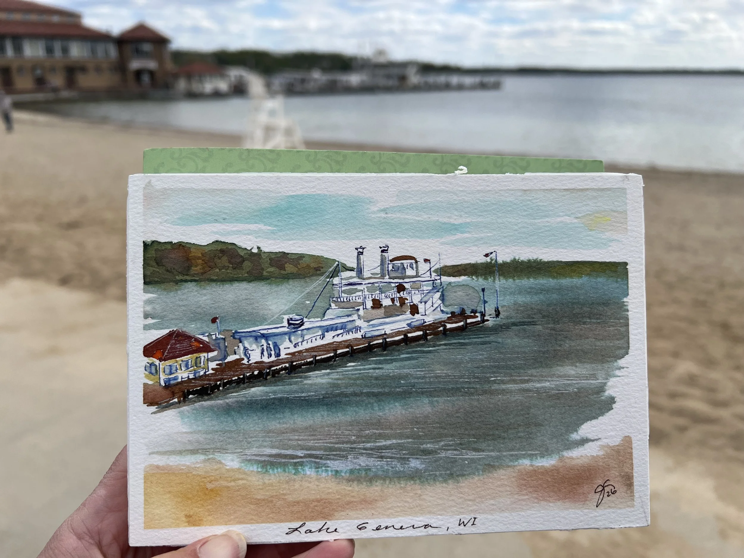 A watercolor painting of a Lake Geneva Boat dock with cruise ships, trees, and a sky with clouds, held in a person's hand against a beach background.