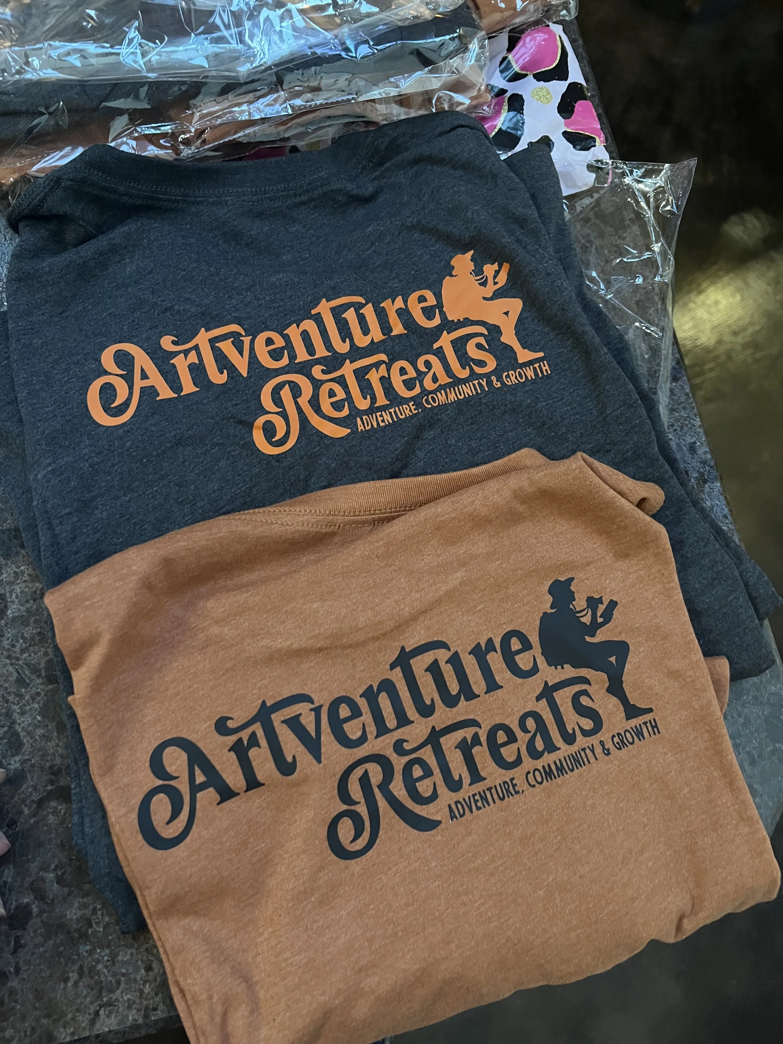 Close-up photo of two folded T-shirts, one dark gray and one brown, with 'Adventure Retreats' printed on the front and a silhouette of a person sitting with a coffee mug.