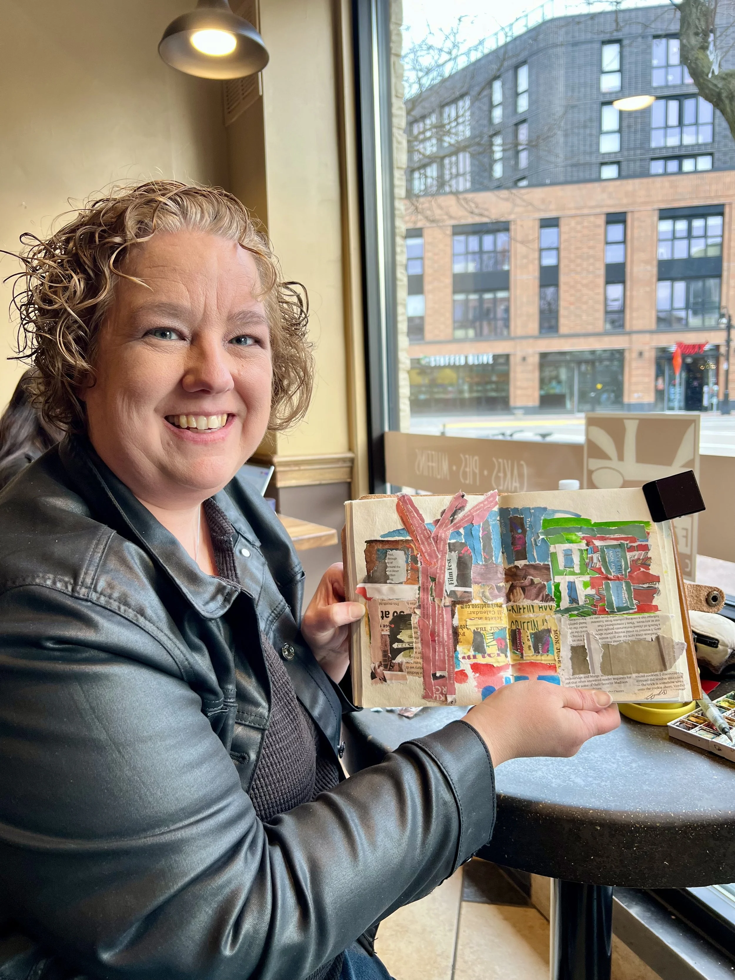 A smiling woman with curly hair wearing a leather jacket sitting at a cafe table, showing a collage of colorful torn paper and magazine clippings in her hand, with large windows and city buildings outside.