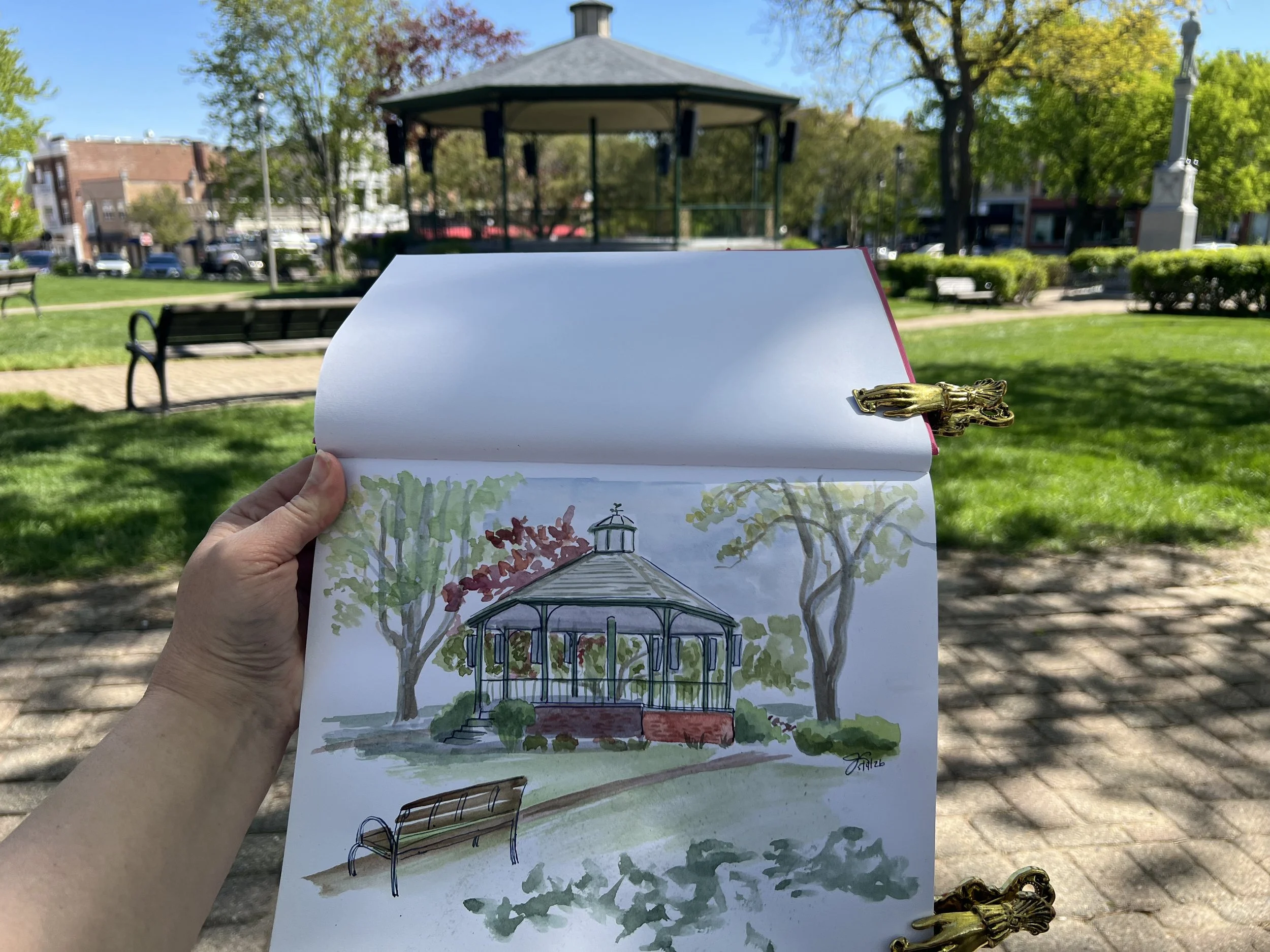 A hand holding an illustrated sketch of a gazebo in a park, with trees and benches, with a pavilion visible in the background under a clear blue sky.