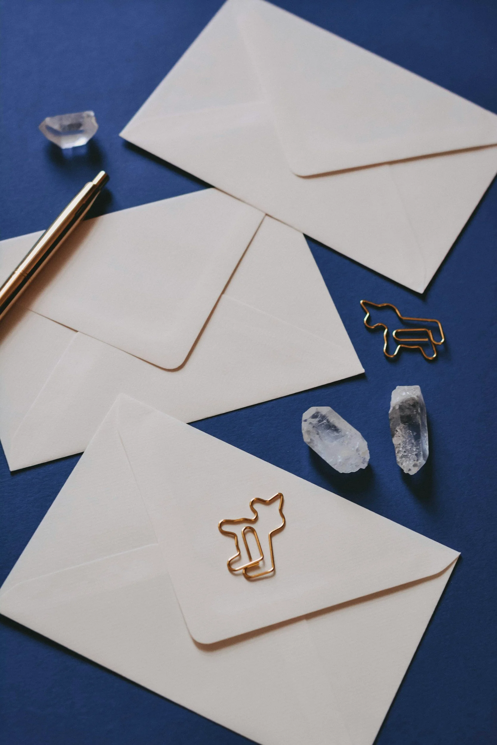 Cream colored envelopes on blue background with gold pen, crystals, and unicorn-shaped paper clips.
