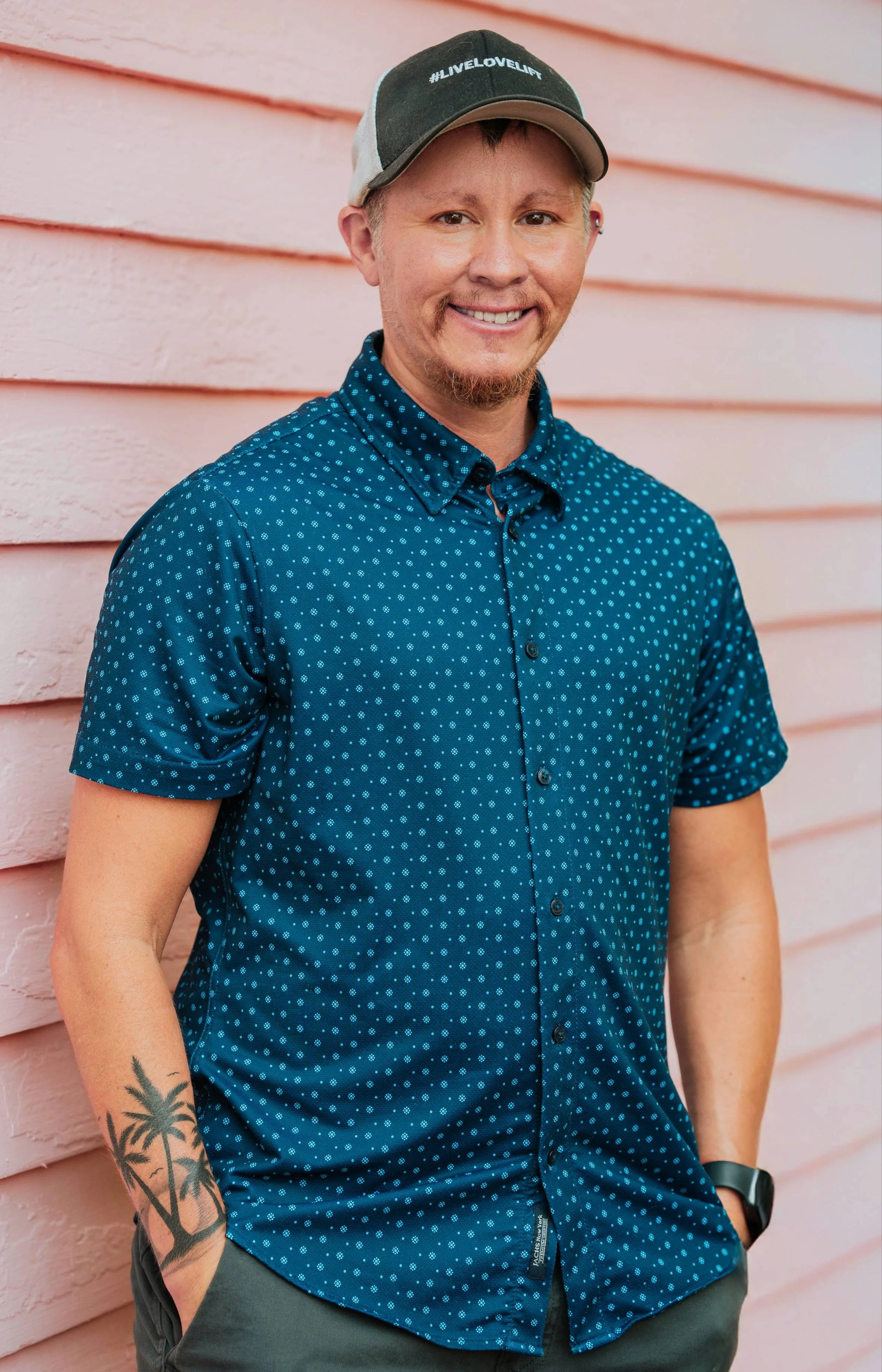 A young man with light skin and a short beard wearing a black and white cap with '#LIVELovelift' written on it, a blue short-sleeved shirt with small white dots, standing against a pink wooden wall. He is smiling and has one hand in his pocket, with a tattoo of palm trees on his forearm.
