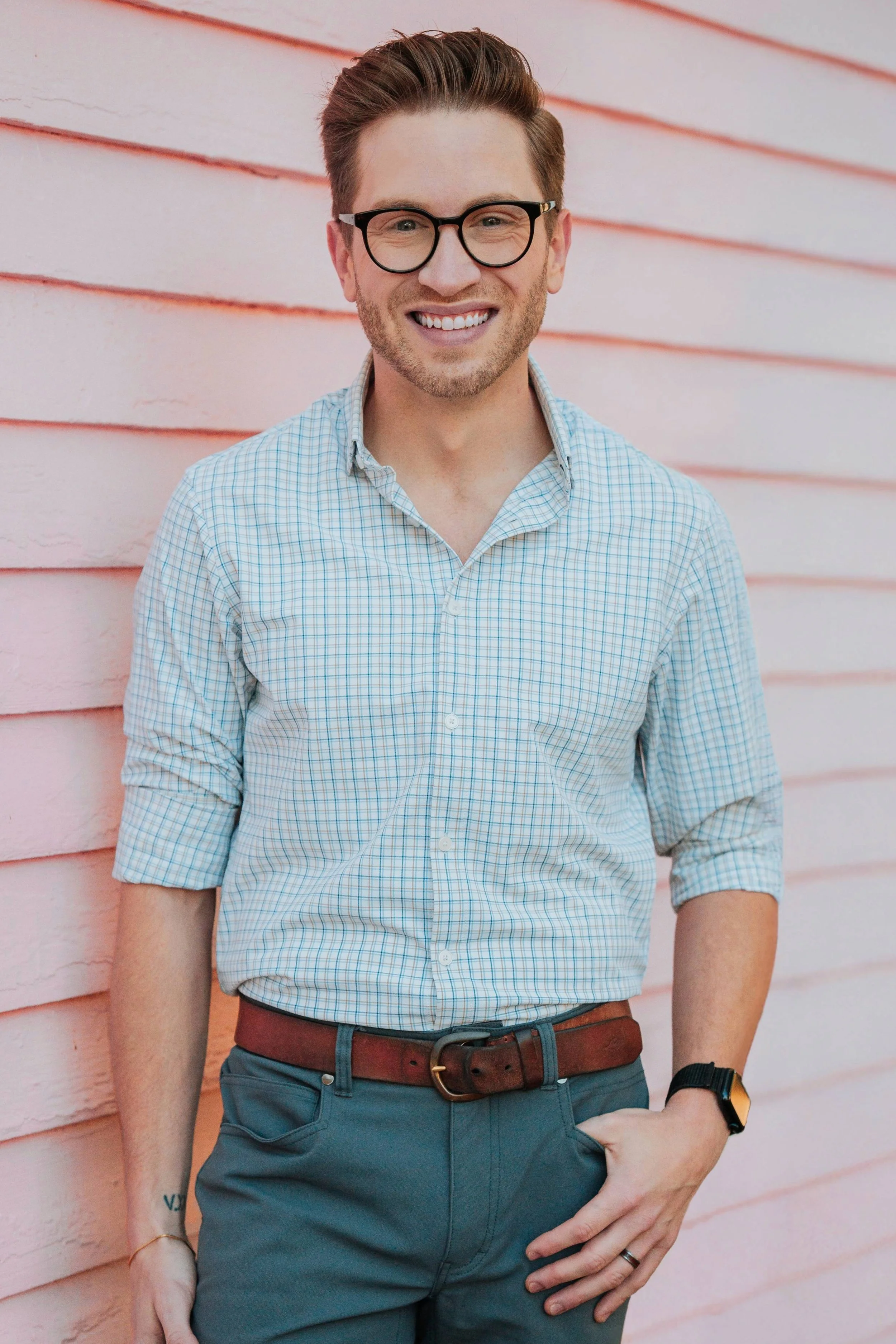 Young man with glasses smiling, standing against a pink wooden wall, wearing a light checkered shirt, dark pants, and a watch.