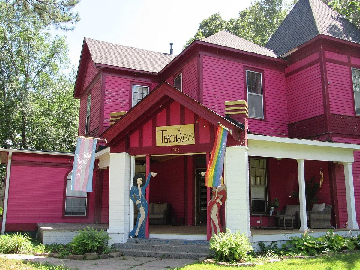 A two-story house painted bright pink with white accents, decorated with pride flags and a welcoming porch with chairs and potted plants, featuring a sign that says 'Teach Love'.