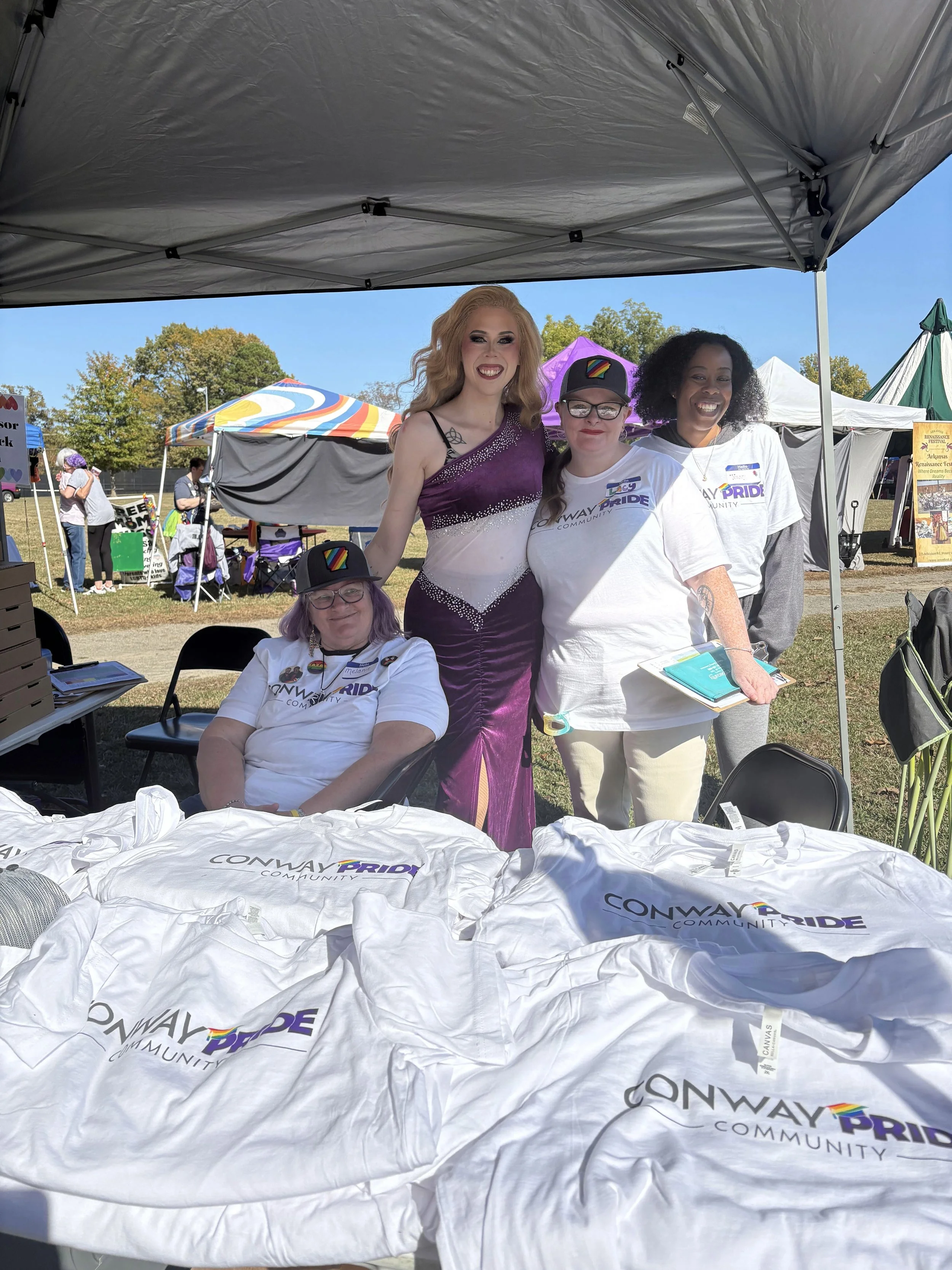 Group of four people standing and sitting under a tent at an outdoor event. The table in the foreground has t-shirts with 'Conway Pride Community' logos. The individuals are smiling, and some wear community event shirts and hats with pride logos.