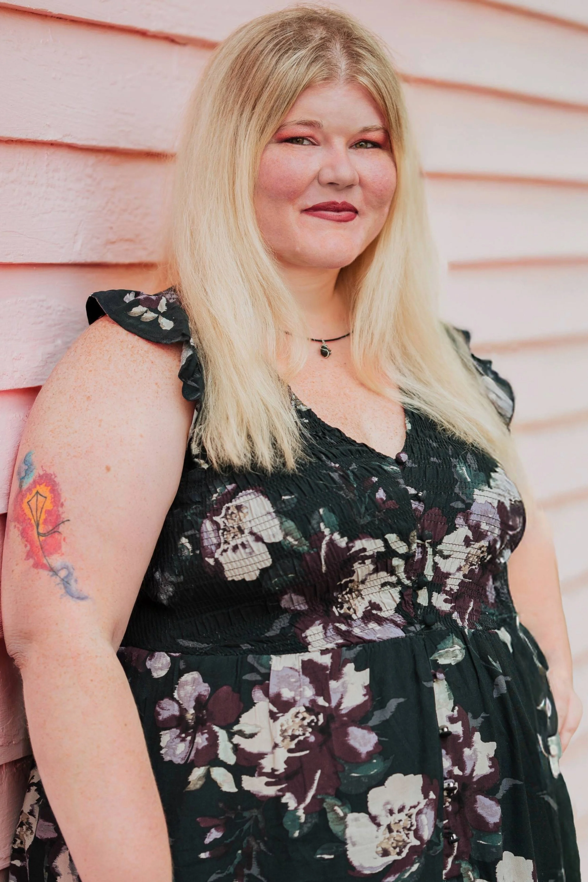 A woman with blonde hair and tattoos on her arm, wearing a black floral dress and a necklace, standing against a pink wooden wall.