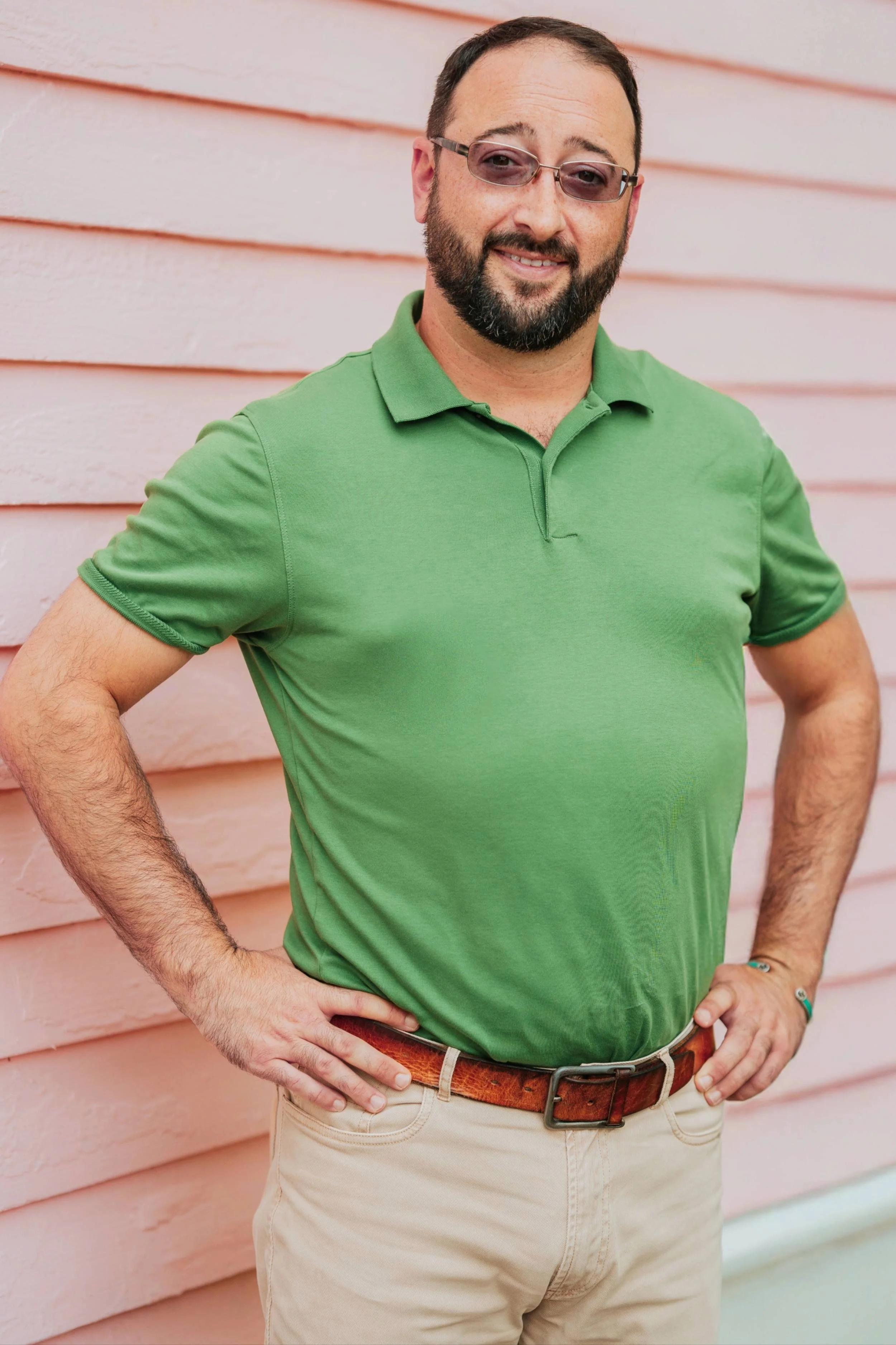A man with a beard and short dark hair, wearing sunglasses, a green polo shirt, and beige pants, standing with his hands on his hips in front of a pink wooden wall.