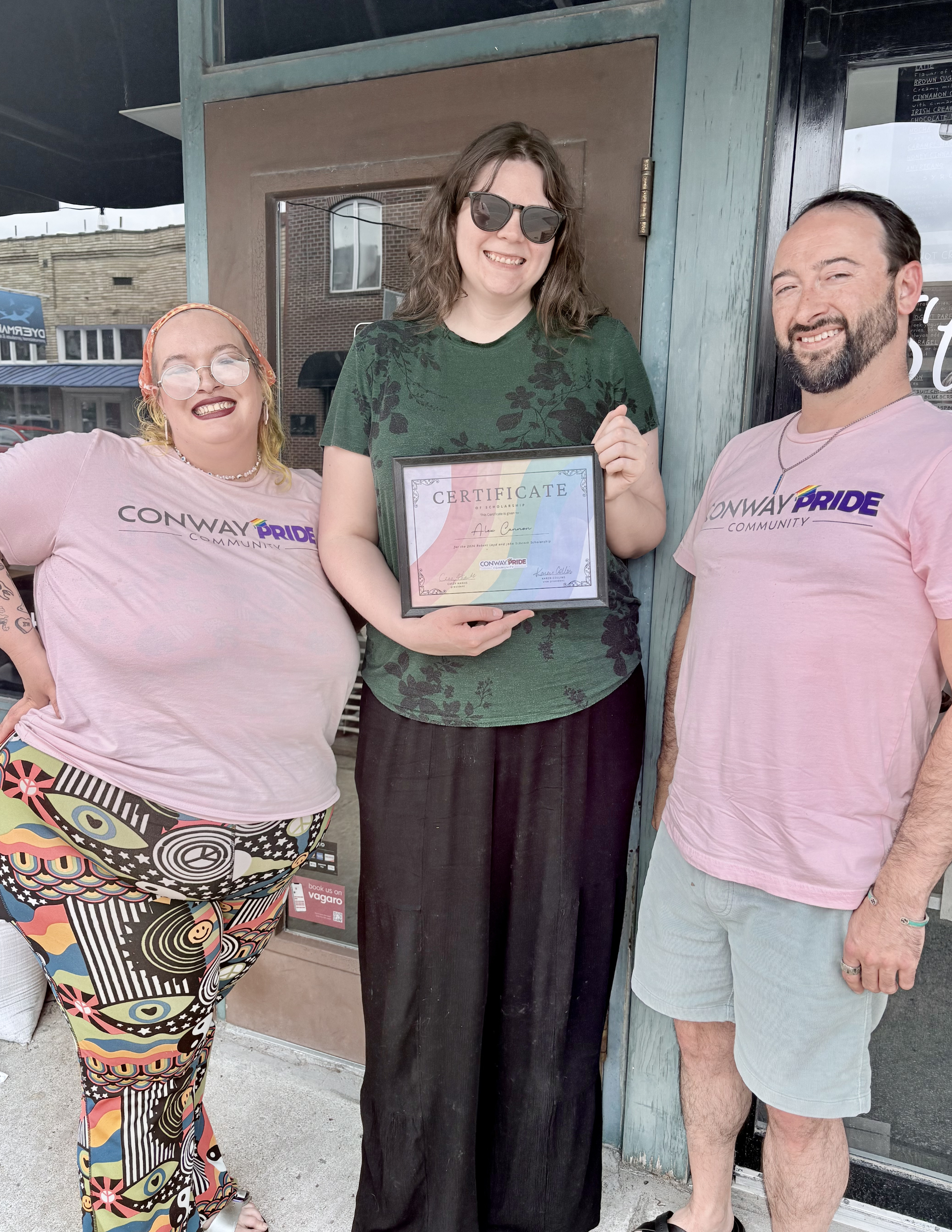 Three people standing outside a store. The person in the middle is holding a certificate and all three are smiling. The person on the left is wearing colorful patterned pants and a pink shirt with 'Conway Pride Community' printed on it. The person on the right is wearing a similar shirt and light-colored shorts. The person in the middle has dark hair, sunglasses, and a green shirt with black floral patterns.