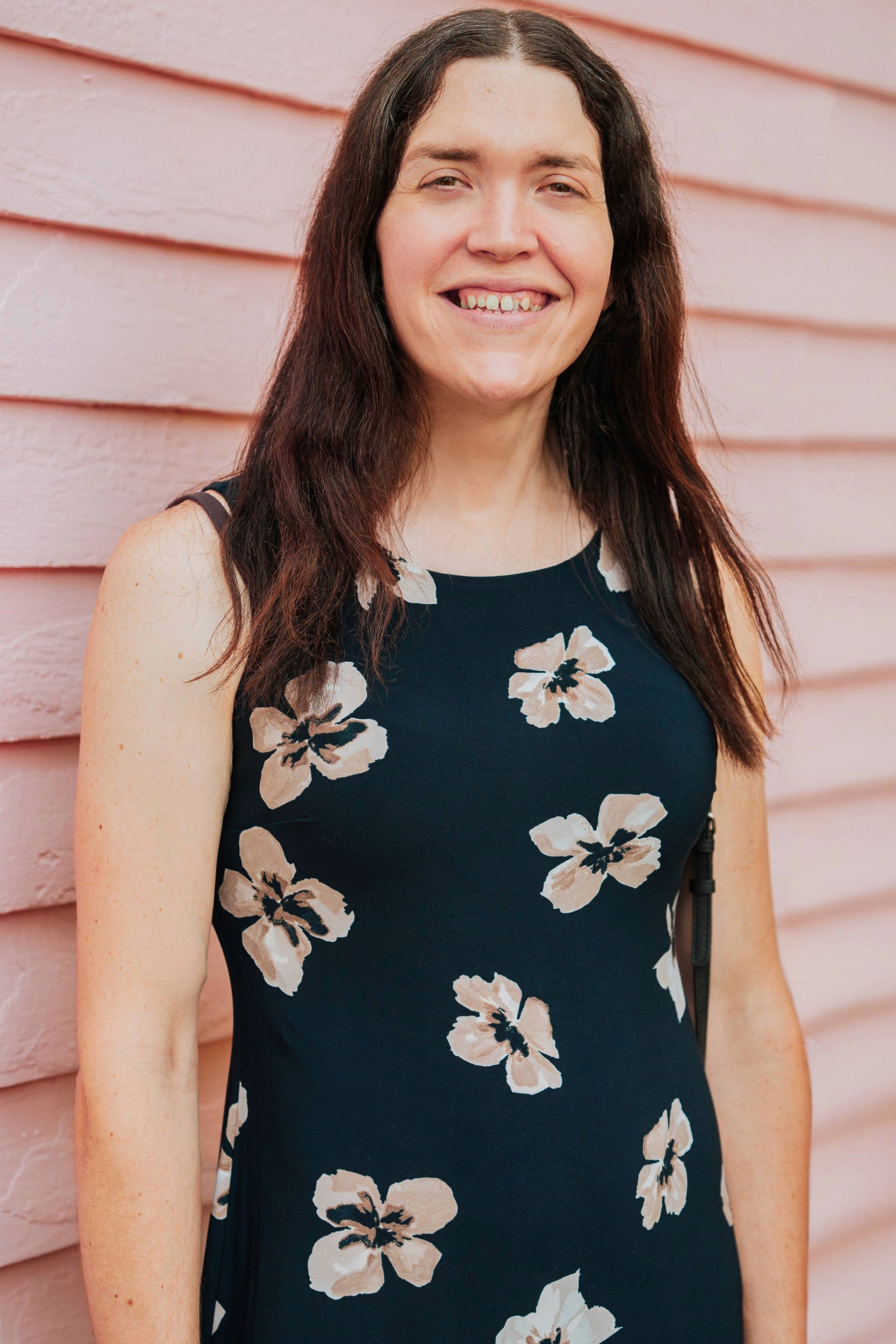 A woman with long brown hair and light skin smiling, wearing a navy blue sleeveless dress with white and beige floral patterns, standing against a pink wooden wall.
