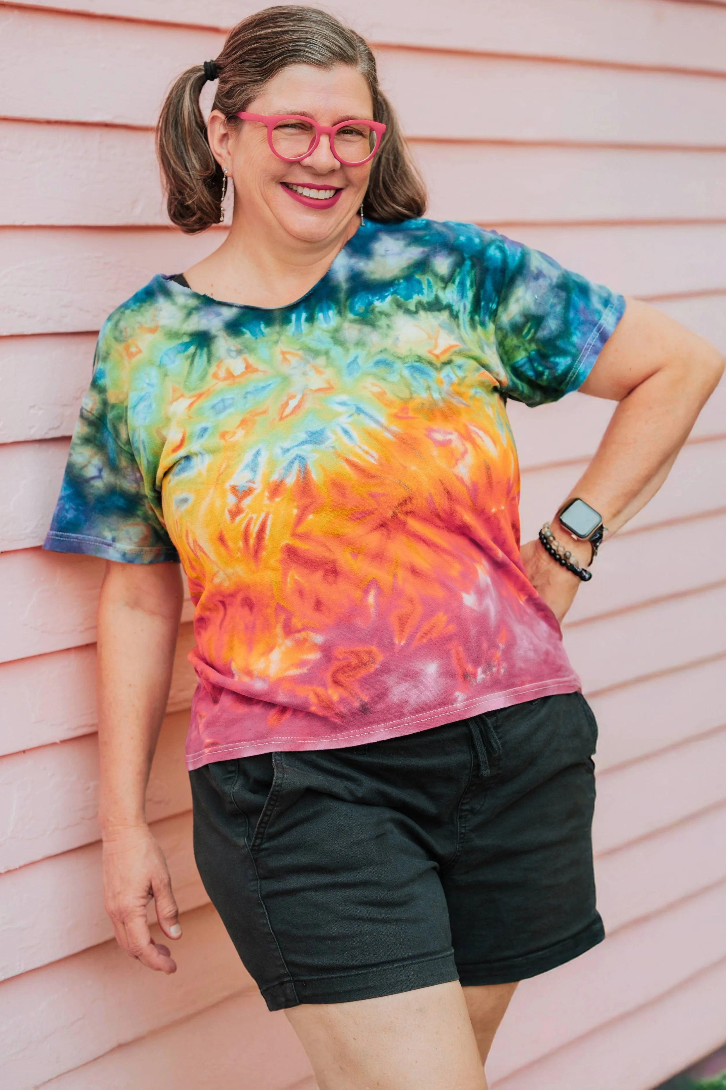 A woman with glasses, earrings, and a watch, wearing a colorful tie-dye T-shirt and black shorts, standing against a pink wooden wall.