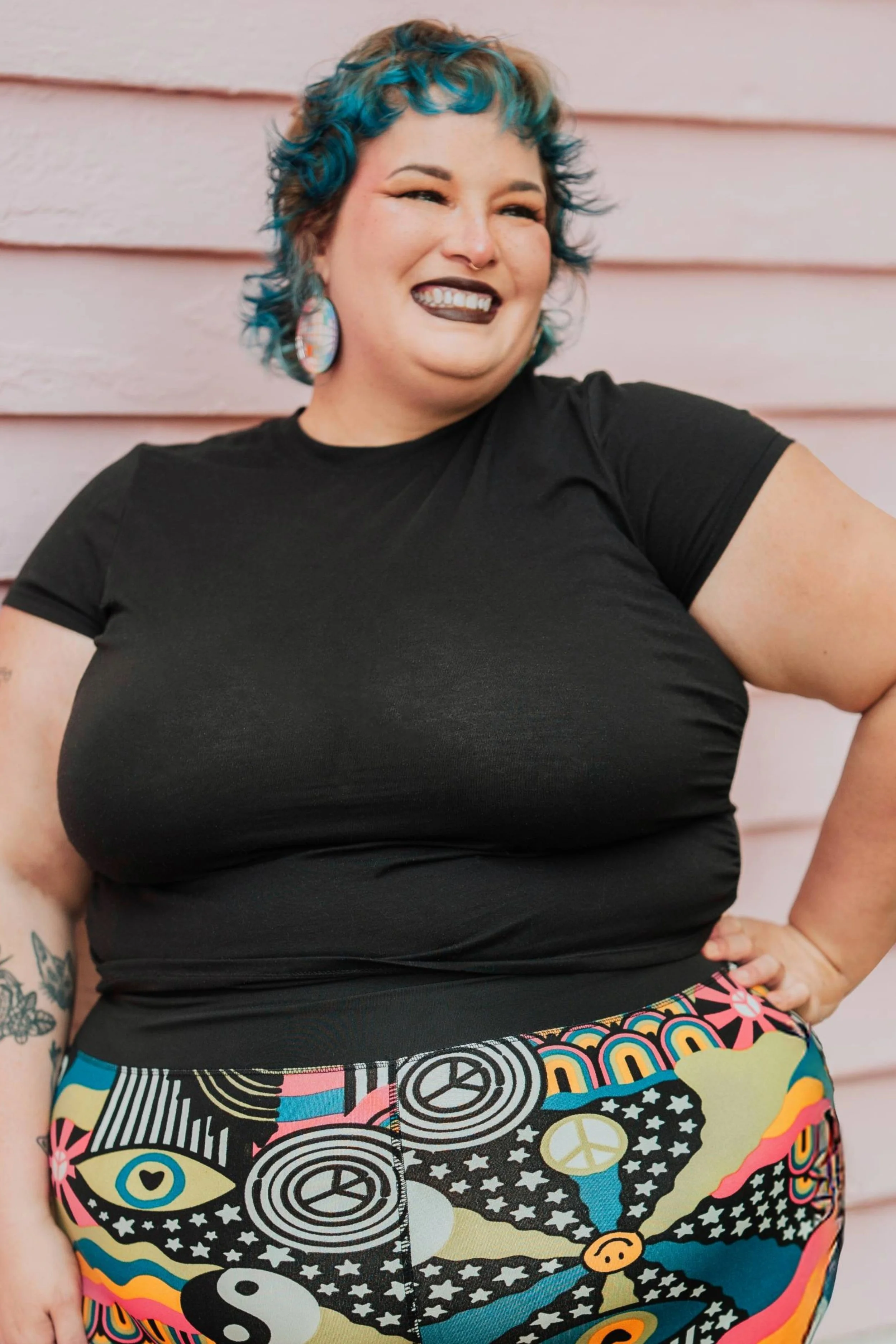 Woman with short curly hair dyed blue and blonde, wearing a black t-shirt and colorful, patterned shorts with symbols like peace signs, yin-yangs, eyes, stars, and smiley faces, standing against a pinkish wall.