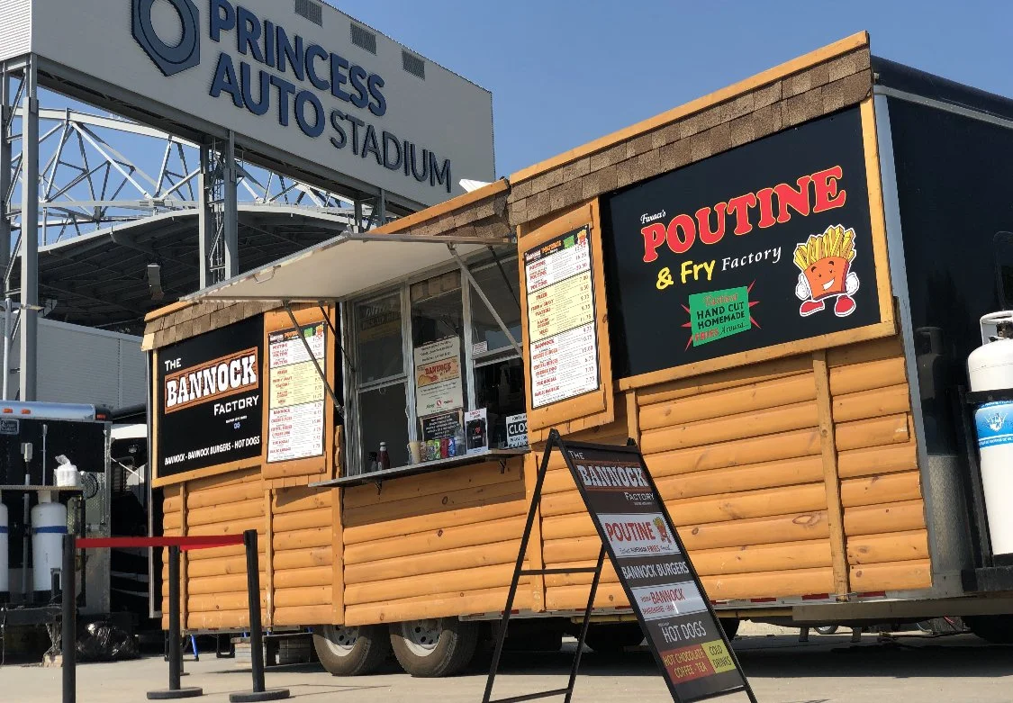 Food truck with signs for The Bannock Factory and Faraci Poutine & Fry Factory, located at Princess Auto Stadium, with a menu and a small standee outside.