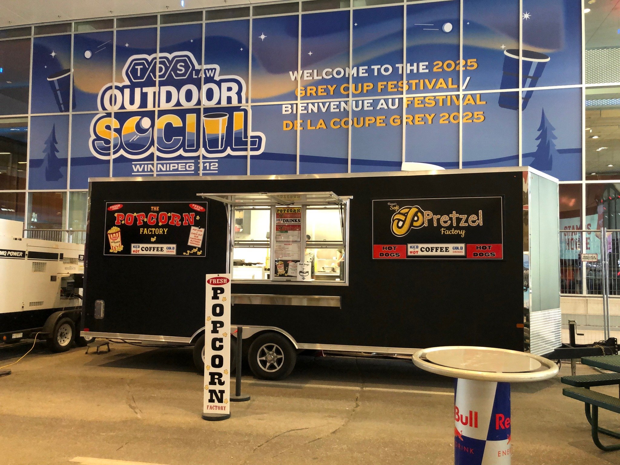 Indoor space with a large blue sign welcoming to the 2025 Grey Cup Festival in both English and French, featuring stars and moon illustrations, and a concession stand selling popcorn, drinks, coffee, hot dogs, and pretzels, with a Red Bull branded table in the foreground.