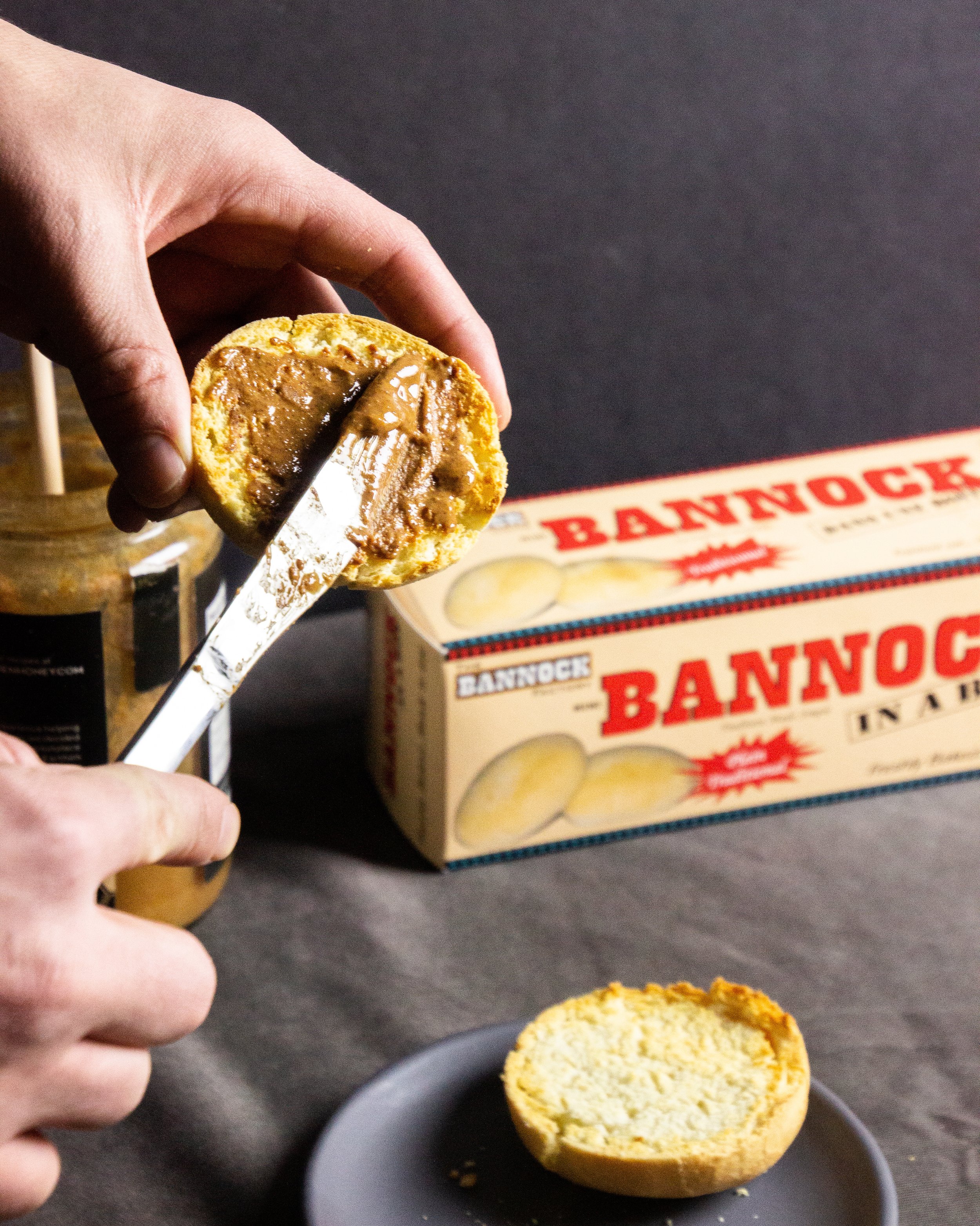 A hand spreading peanut butter on the bottom half of a toasted biscuit, with a box of 'Bannock' crackers and a small black plate in the background.
