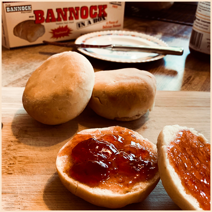 Two bread rolls with jelly, two plain bread rolls, a plate with a knife, a box of Bannock in a Box, and a paper plate on a wooden surface.