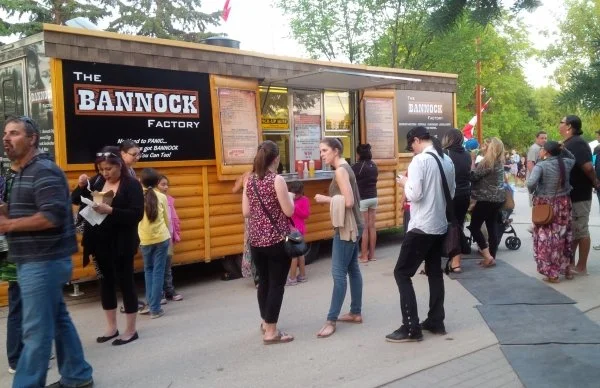 People standing in line at a food stand called 'The Bannock Factory' during daytime, with trees in the background and a sidewalk in front.