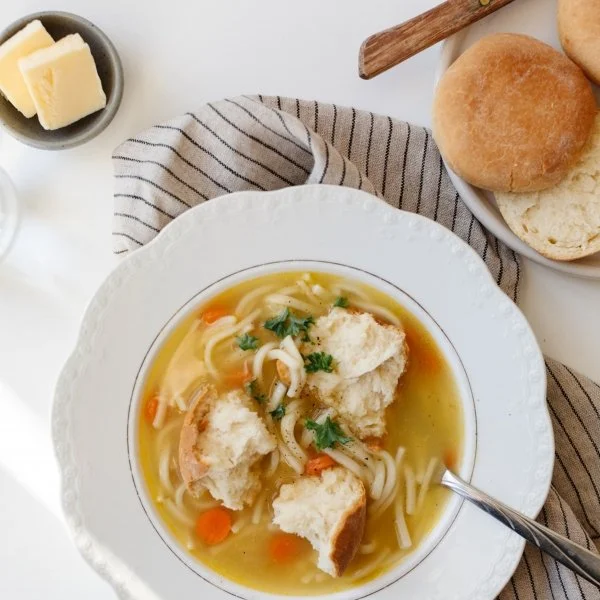 A bowl of chicken noodle soup with bannock bread pieces, a small dish of butter, and bread rolls on a white table with a striped cloth.