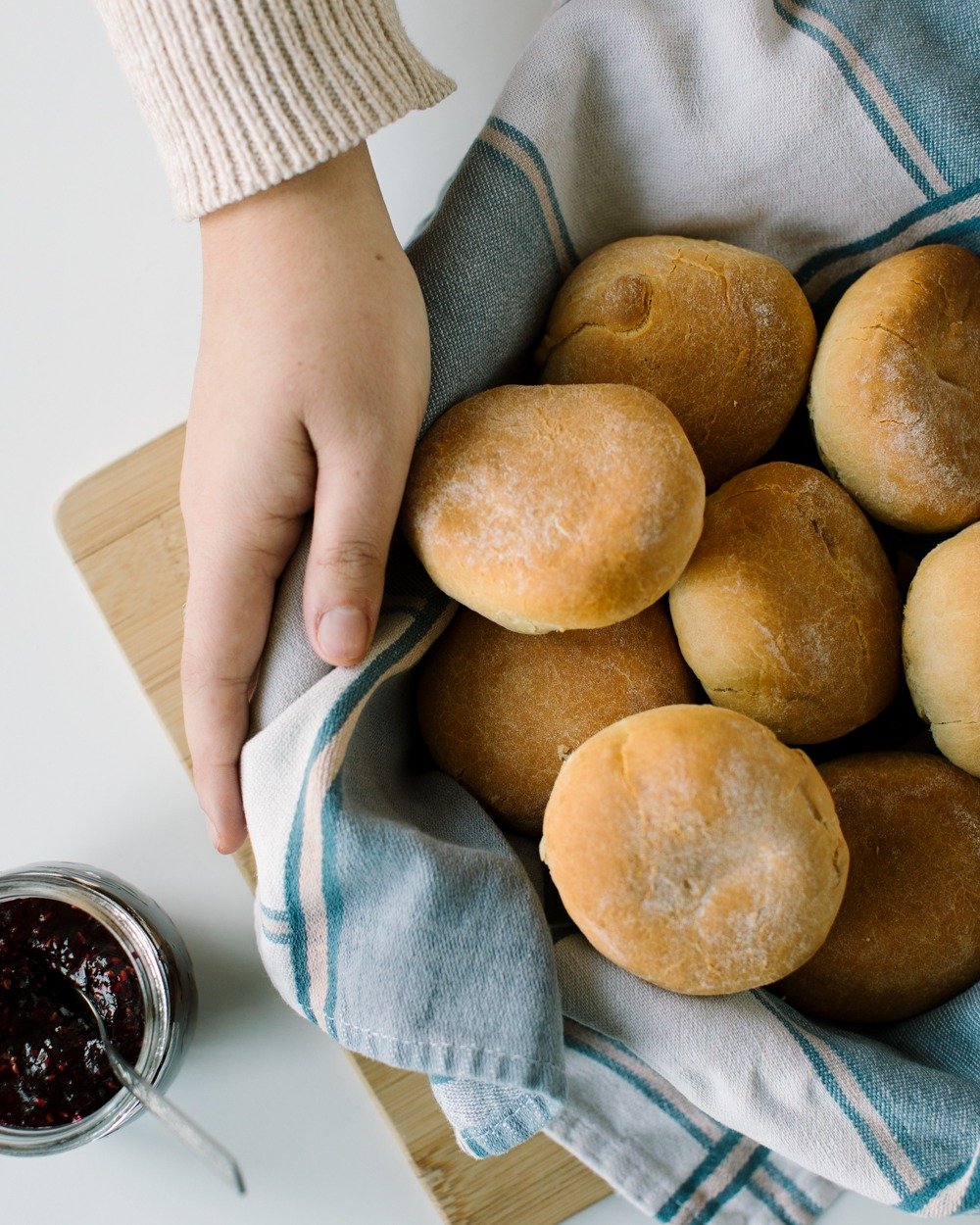 A hand holding a blue and white striped cloth basket filled with round bread rolls, with a jar of dark berry jam nearby.