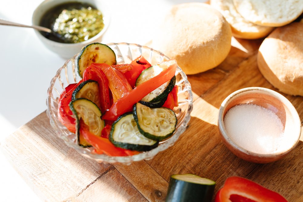 A glass bowl of sliced cucumbers, red bell peppers, and tomatoes on a wooden surface, with a small bowl of salt, a plate of bannock buns, and a small dish of green sauce nearby.