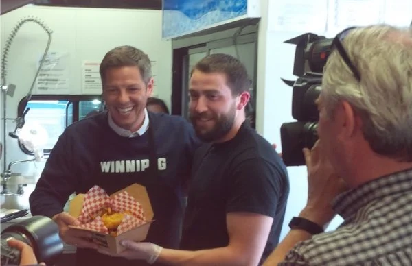 Mayor Brian Bowman smiling at a camera, with Anthony Faraci. one holding a tray of food, in a kitchen or food prep area.