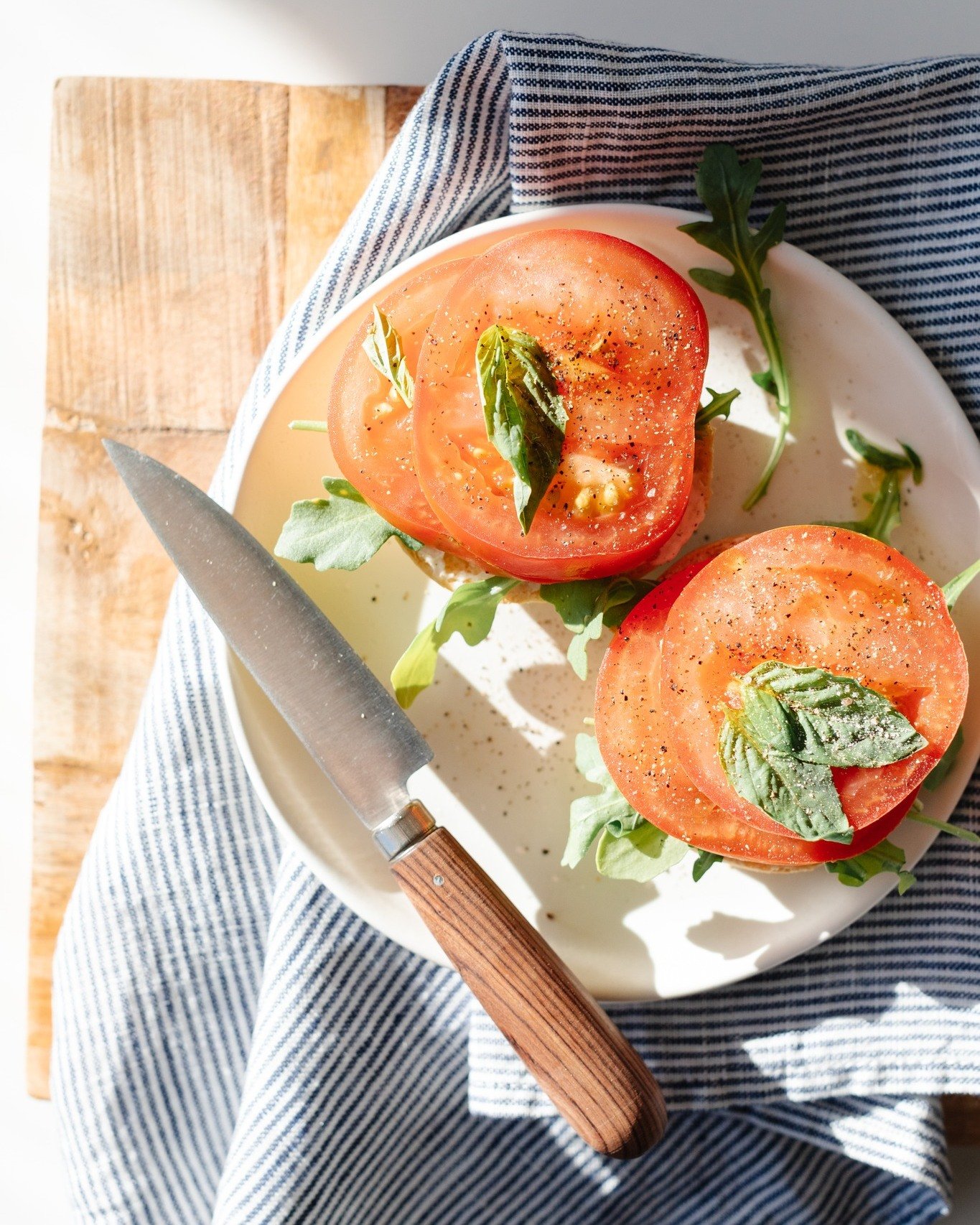 Fresh tomato slices on a plate garnished with basil leaves, sprinkled with black pepper, with a knife on a blue-striped cloth.
