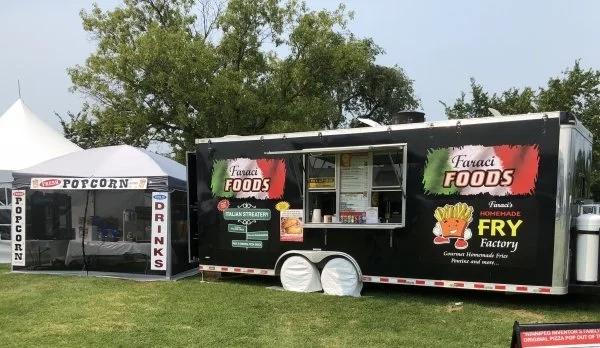A black food truck with green and red branding, serving Italian street food and fries, parked on grass with trees in the background, and a white tent selling popcorn and drinks nearby.