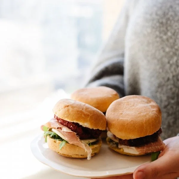 Person holding a white plate with three mini bannock sandwiches containing various deli meats, greens, and condiments.