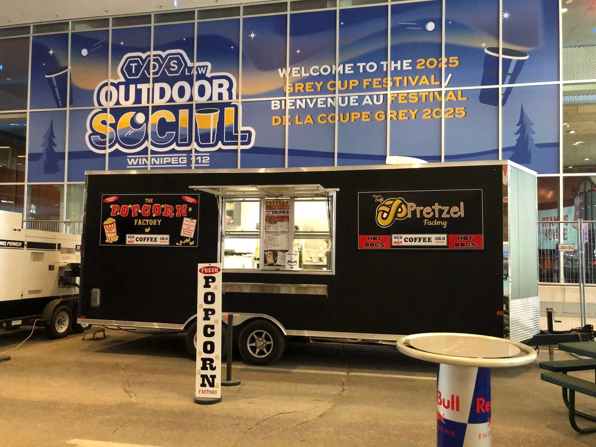 Large digital sign welcoming visitors to the 2025 Grey Cup Festival at Winnipeg, with logos, starry sky, and stylized trees. A concession stand in front offers popcorn, coffee, hot dogs, and drinks.
