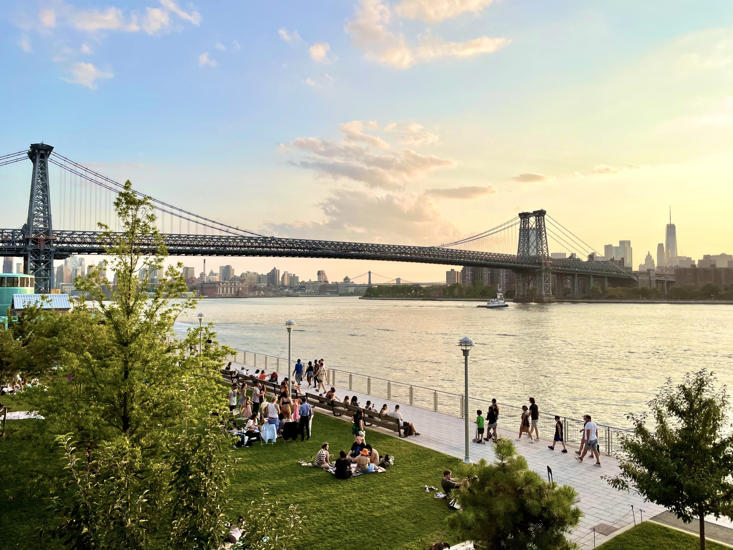 People sitting and walking along a riverside park in New York City, with the Brooklyn Bridge and Manhattan skyline in the background during sunset.