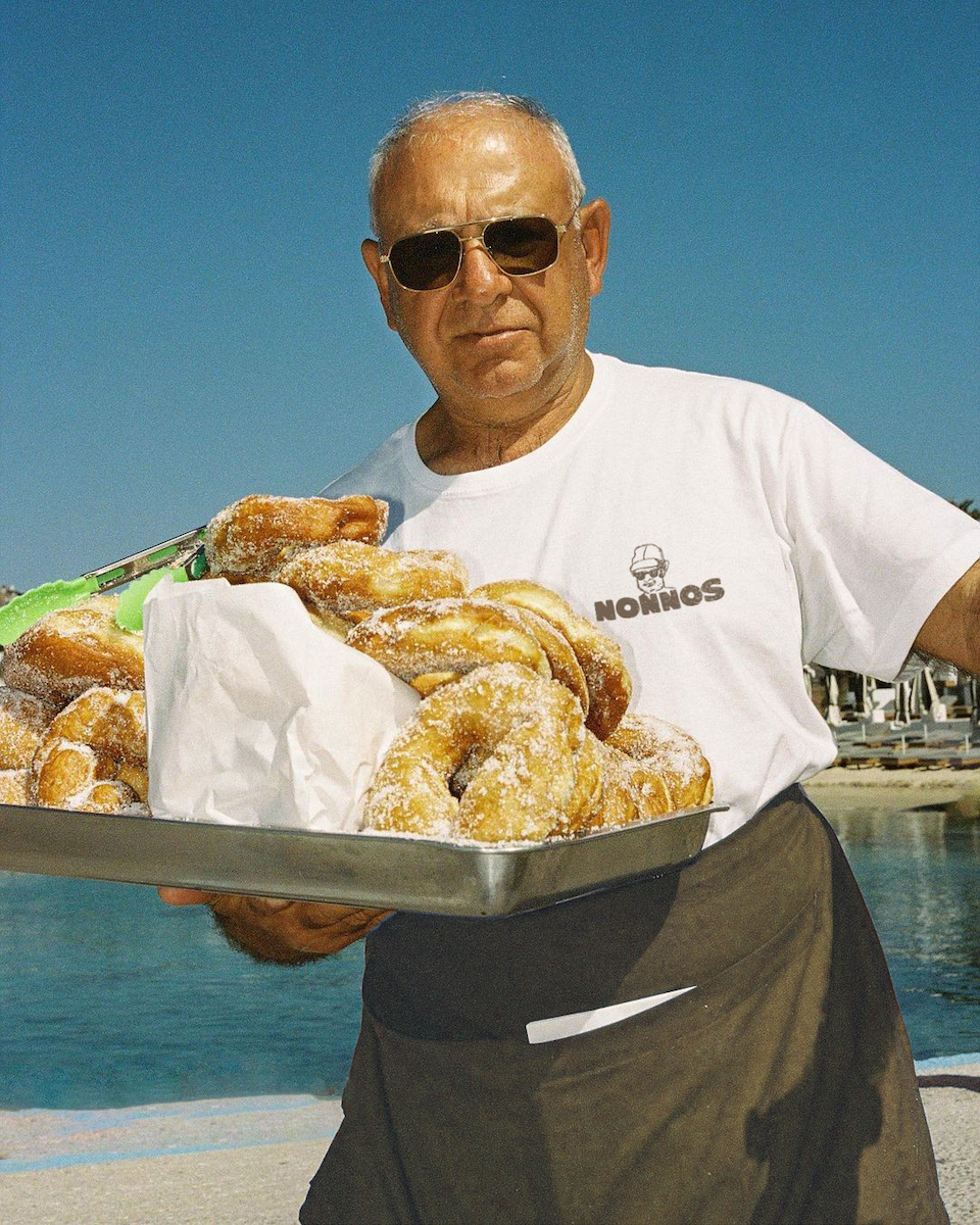 An elderly man wearing sunglasses and a white T-shirt holding a tray of powdered donuts by a poolside.