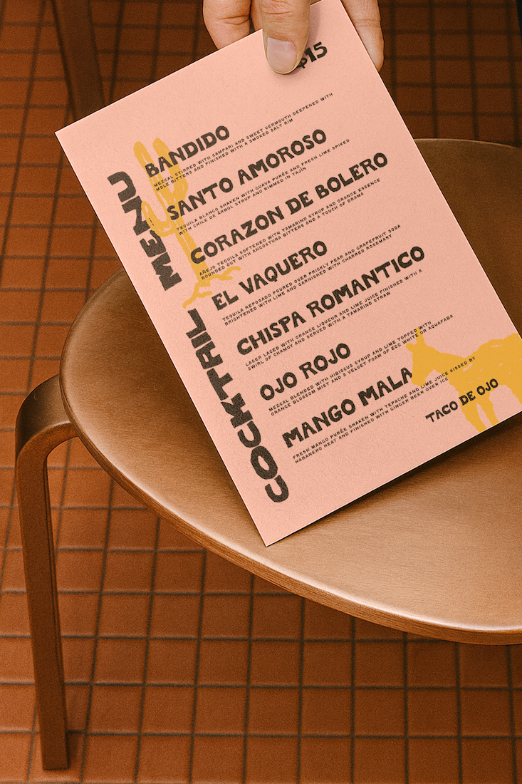 A hand holding a pink menu with black and yellow text on a wooden chair with a tiled orange floor.
