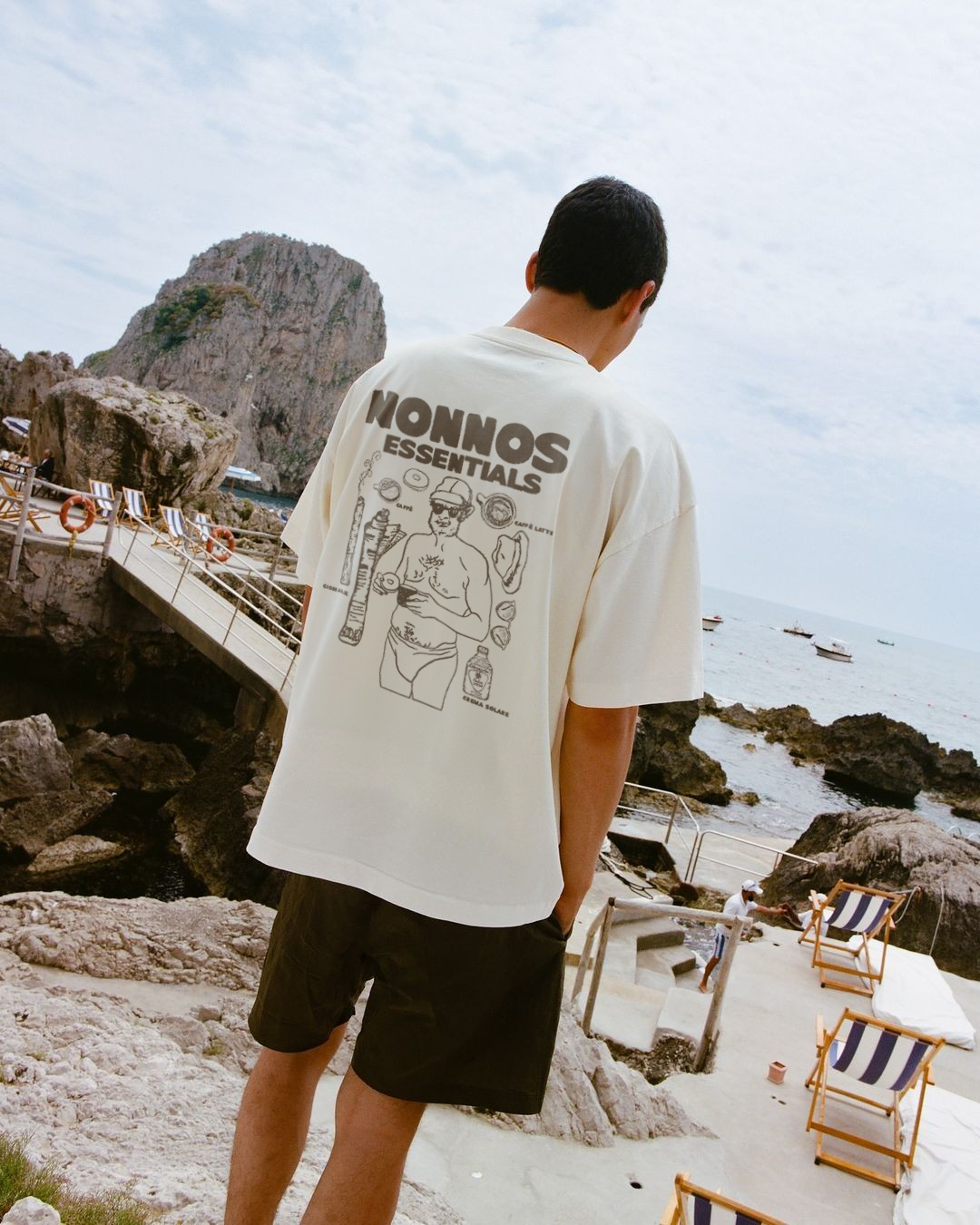 A man wearing a white t-shirt with graphic designs and the words 'NONNOS ESSENTIALS' stands near rocky coastline on a sunny day, looking out toward the sea with boats in the distance and a rock formation in the background.