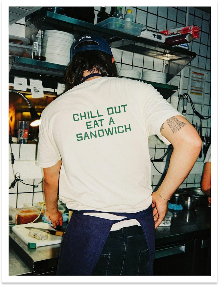 A person with dark hair wearing a white t-shirt with green text that reads 'Chill Out Eat a Sandwich', a blue cap, and dark jeans, working in a kitchen with shelves of dishes and cooking supplies in the background.