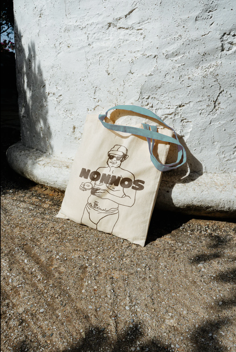 A tote bag with a drawing of a woman in sunglasses and a hat, holding a phone, leaning against a white textured wall on a sandy ground.