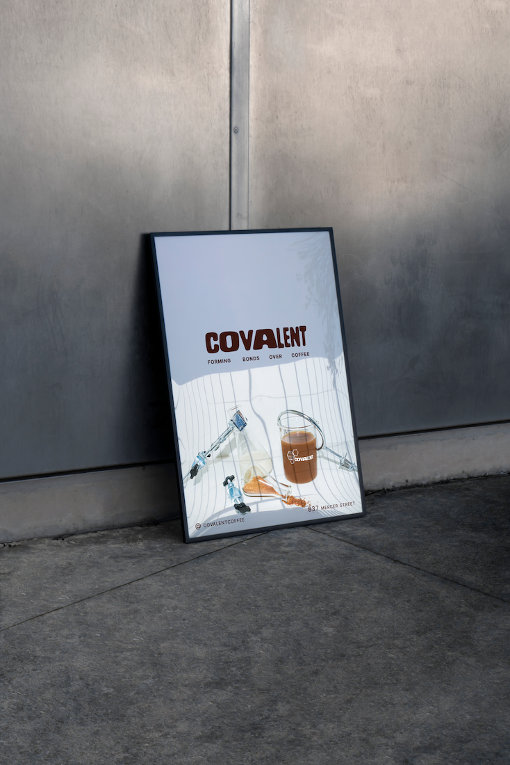 Framed poster on sidewalk featuring the word 'Covalent' with the tagline 'Forming Bonds Over Coffee' and an illustration of graduated cylinders, a liquid, and coffee cup branding.