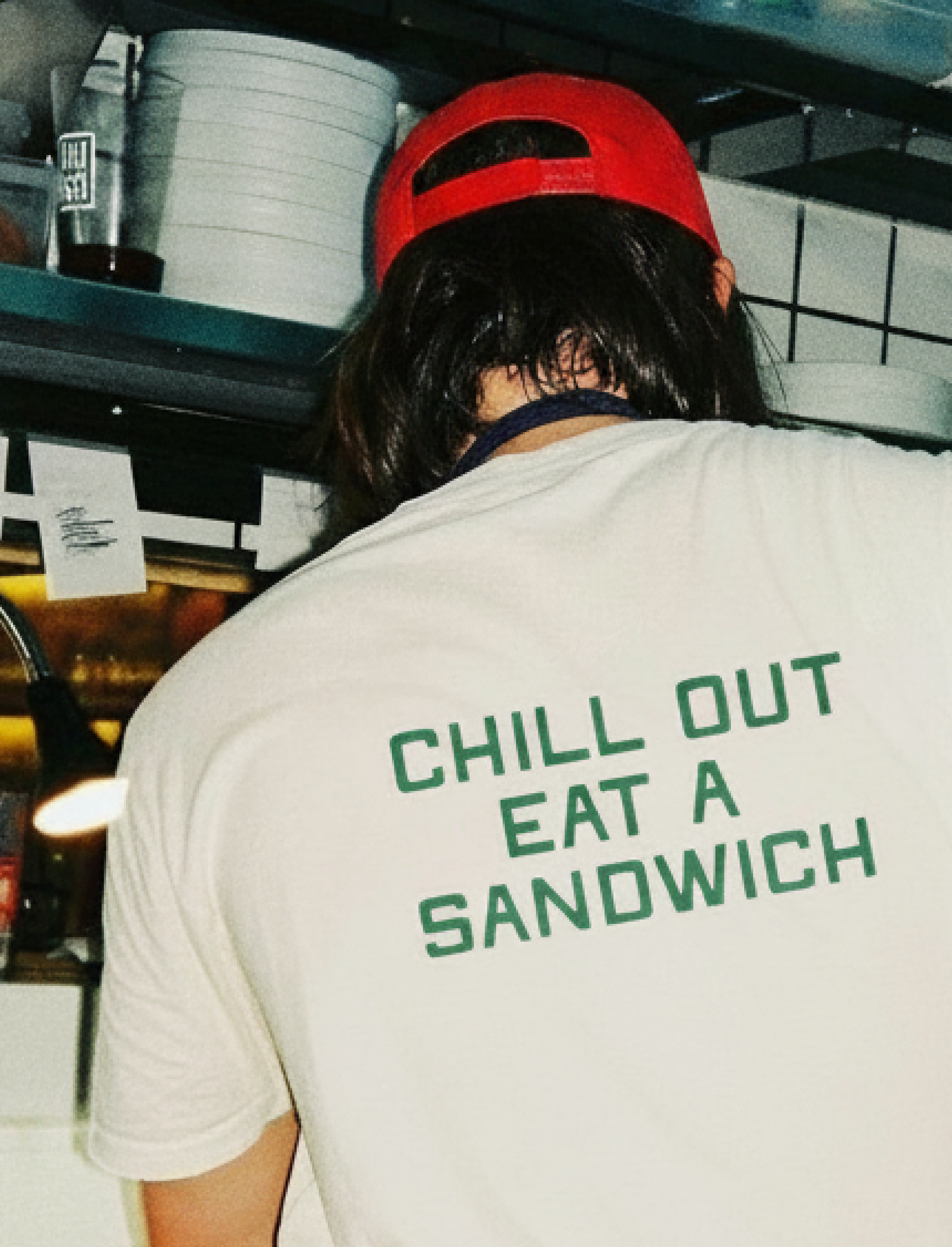 Person wearing a white T-shirt with green text, a red cap, and dark hair, in a kitchen setting with dishes and containers on shelves.